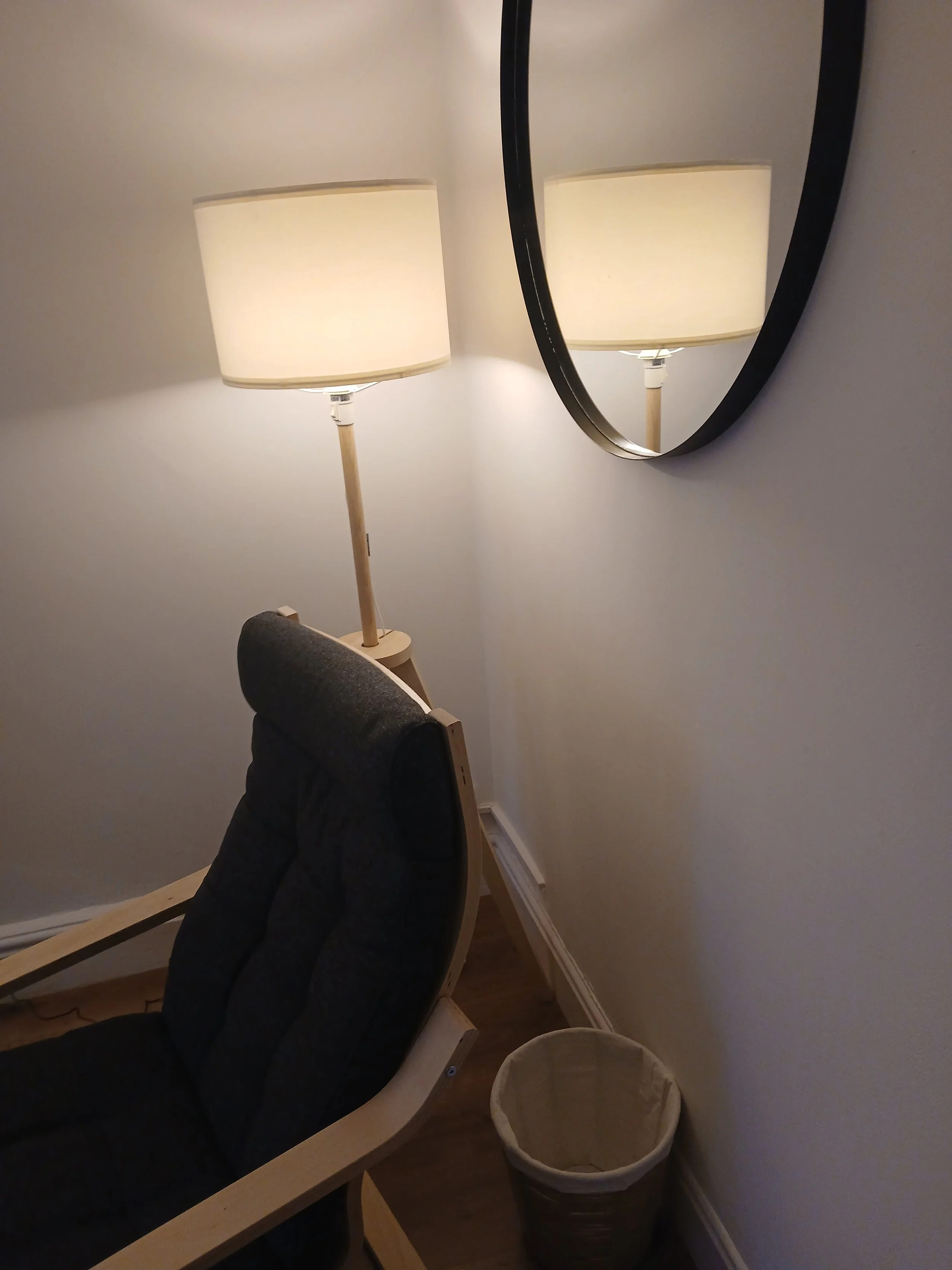 A cozy corner featuring a standing lamp with a white shade, a black upholstered chair with a wooden frame, a round wall mirror, and a small beige trash bin beneath the mirror on a wooden floor.