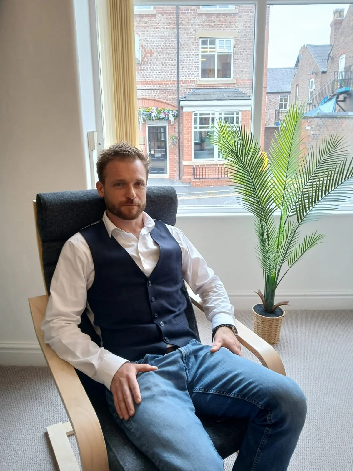 A man with light skin, short brown hair, and a beard sitting comfortably in a black office chair in front of a window with a view of brick buildings outside. He is wearing a white shirt, a dark vest, and blue jeans, with one hand resting on his lap and the other on the armrest. There is a large potted palm plant to his right.