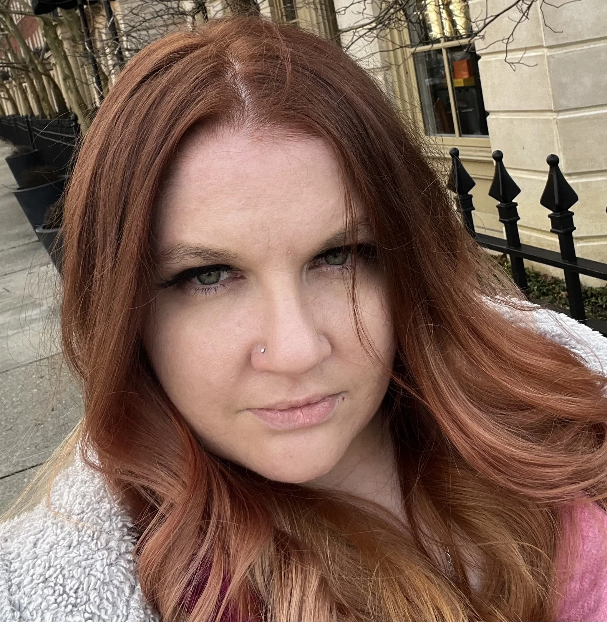 Close-up of a woman with red hair and a small piercing on her nose, outdoors with trees and a building with large windows and a black fence in the background.