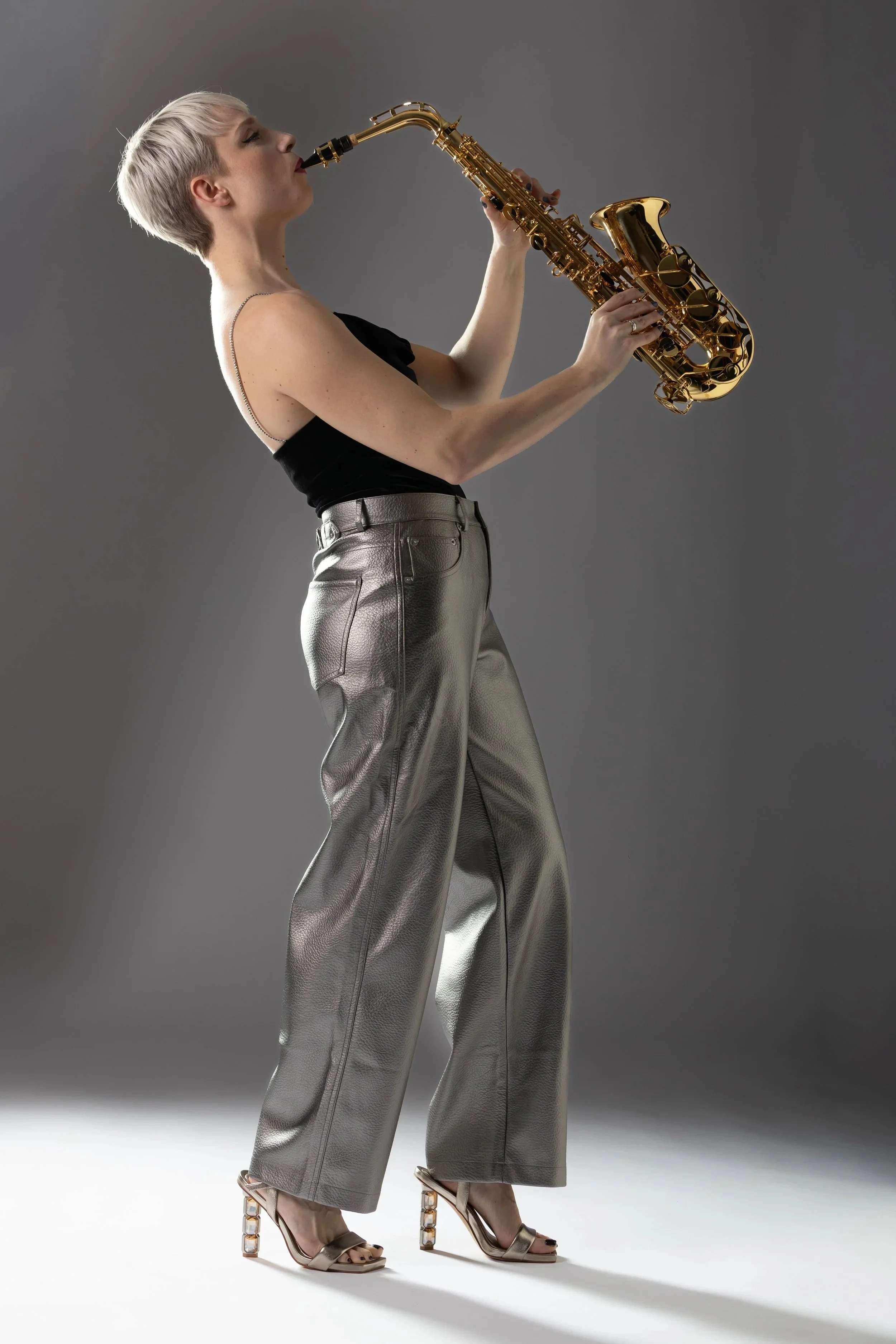 A woman with short blonde hair playing a saxophone against a gray background, wearing a black top, metallic silver wide-leg pants, and high heels.