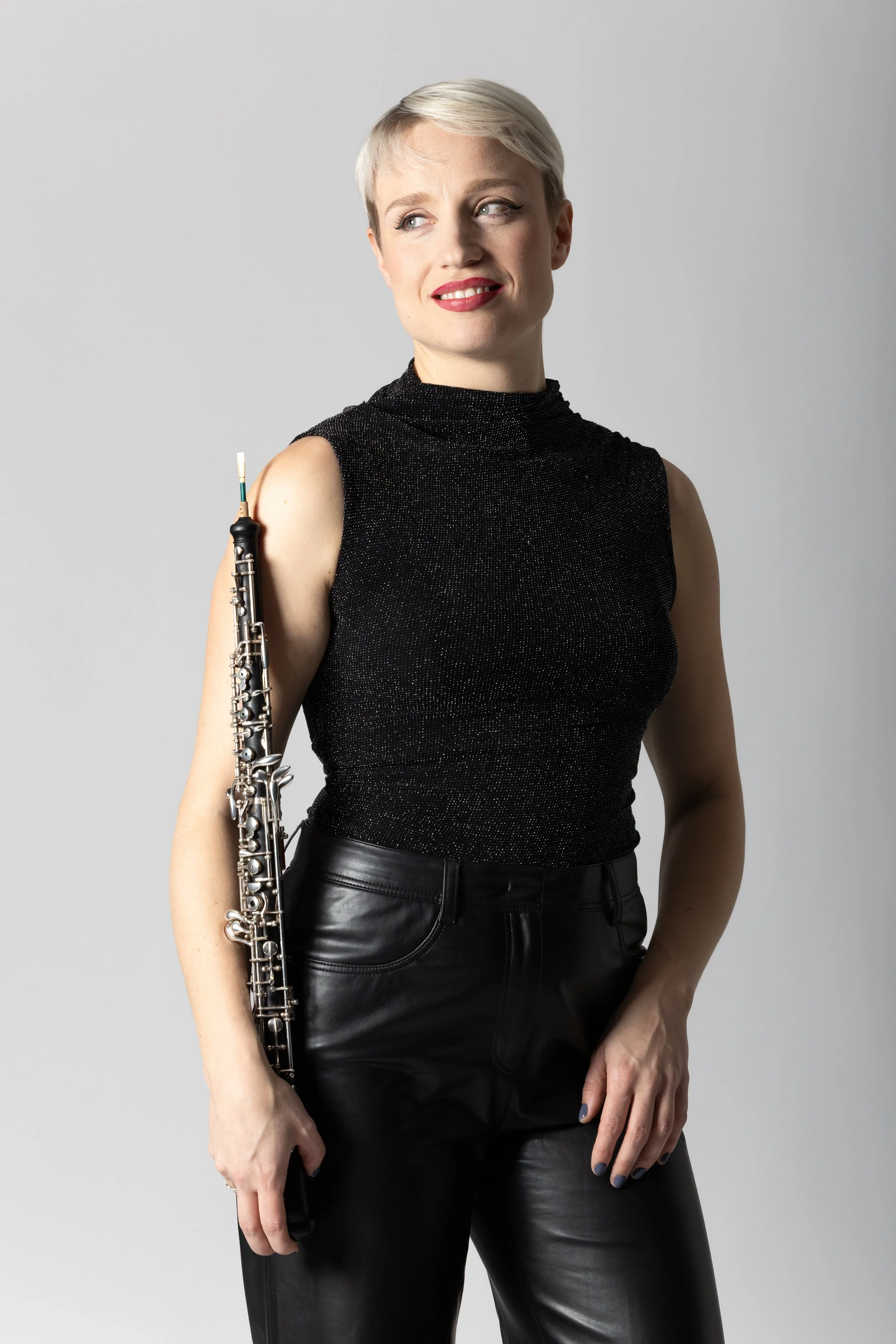 A woman with short blonde hair holding a clarinet, wearing a black sleeveless top and black leather pants, standing against a plain gray background.