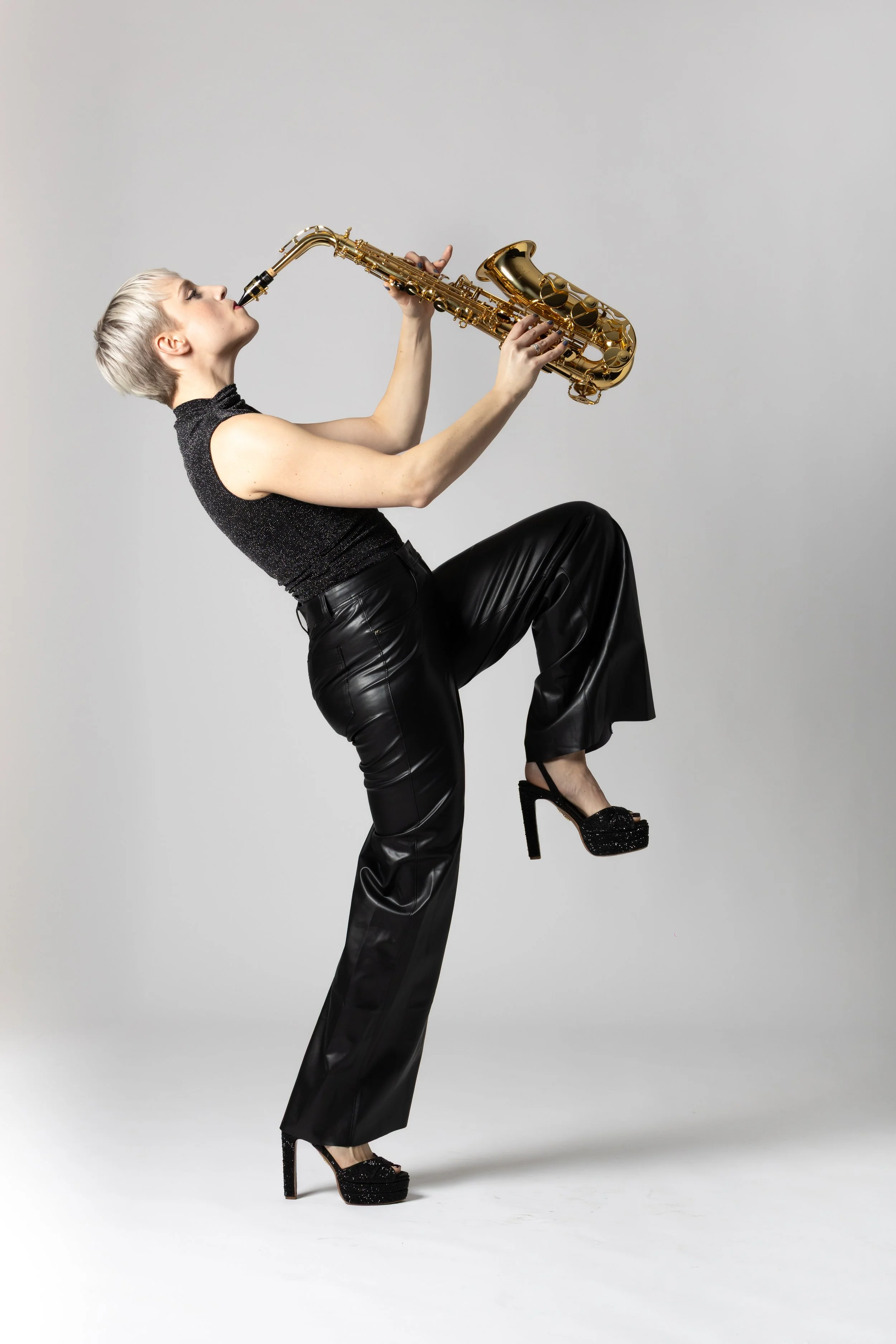 A woman with short blonde hair wearing a black sleeveless top and black leather pants, standing with one leg raised, playing a gold saxophone against a plain light gray background.