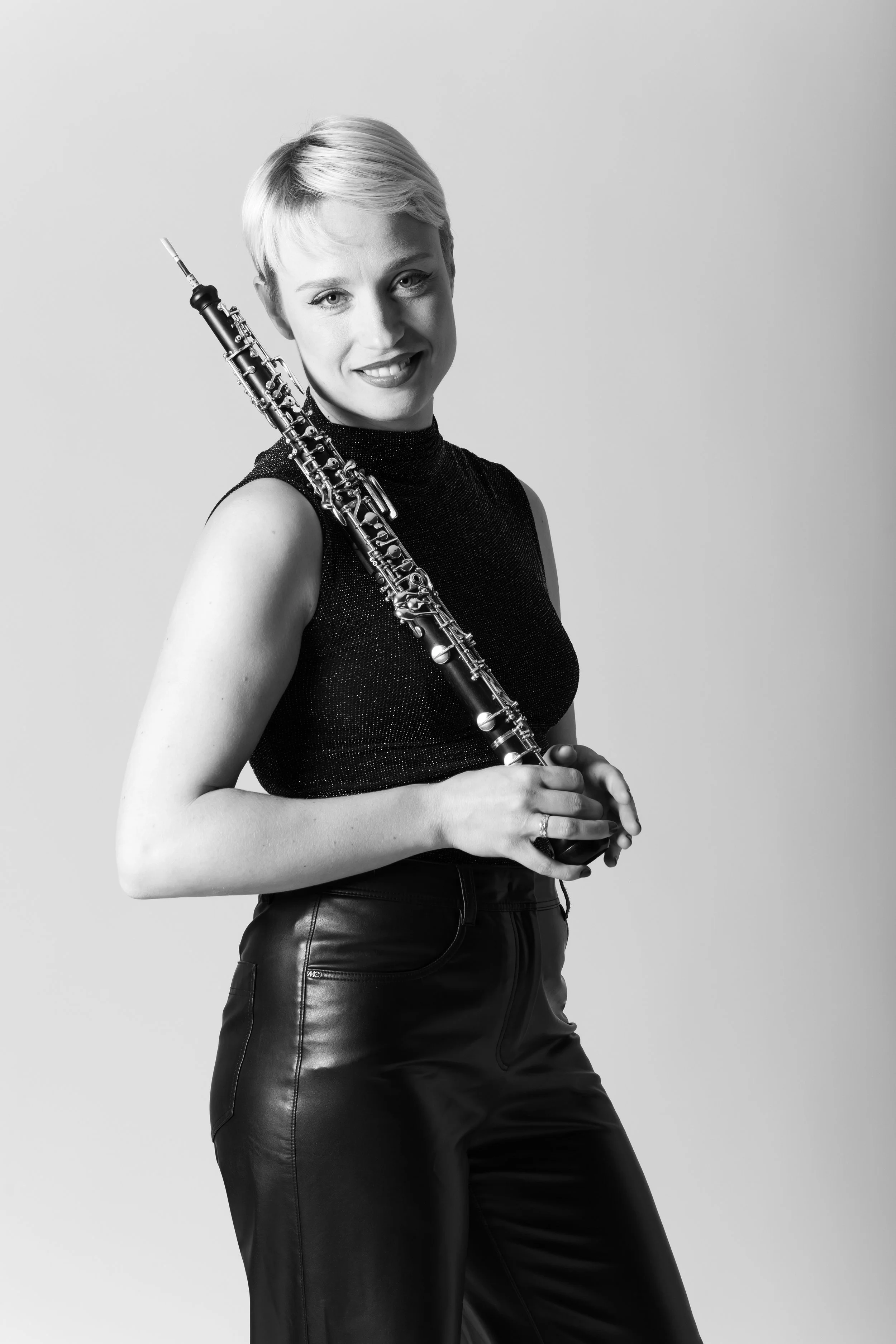 Black and white photo of a woman with short blonde hair, smiling, holding a flute across her shoulder, wearing a sleeveless top and leather pants.
