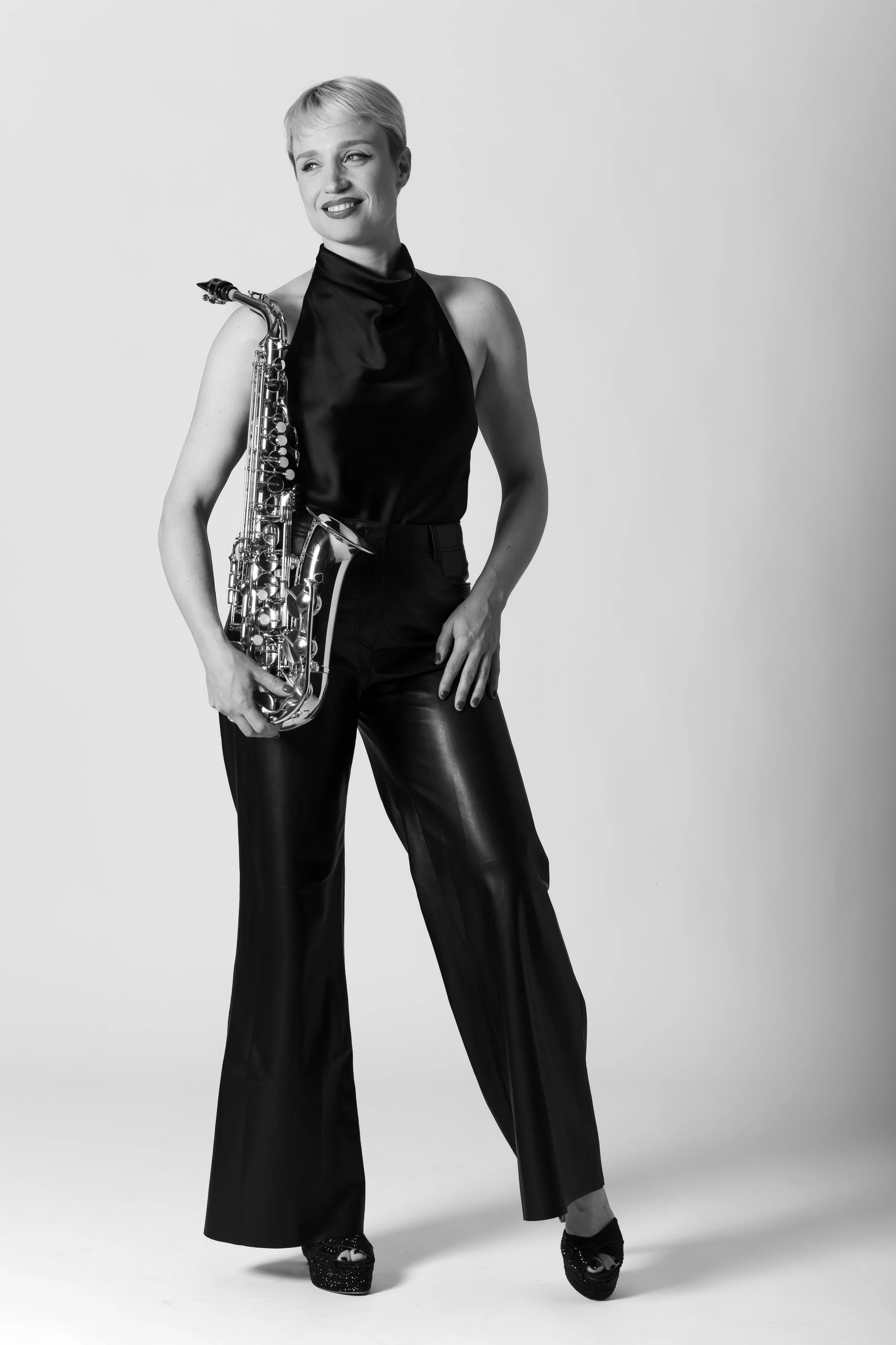 Black and white photo of a woman holding a saxophone, dressed in shiny black pants and a sleeveless top, standing against a plain background.