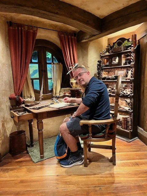A man sitting at a wooden desk in a cozy room with wooden beams, a window with red curtains, and shelves filled with books and decorative items.