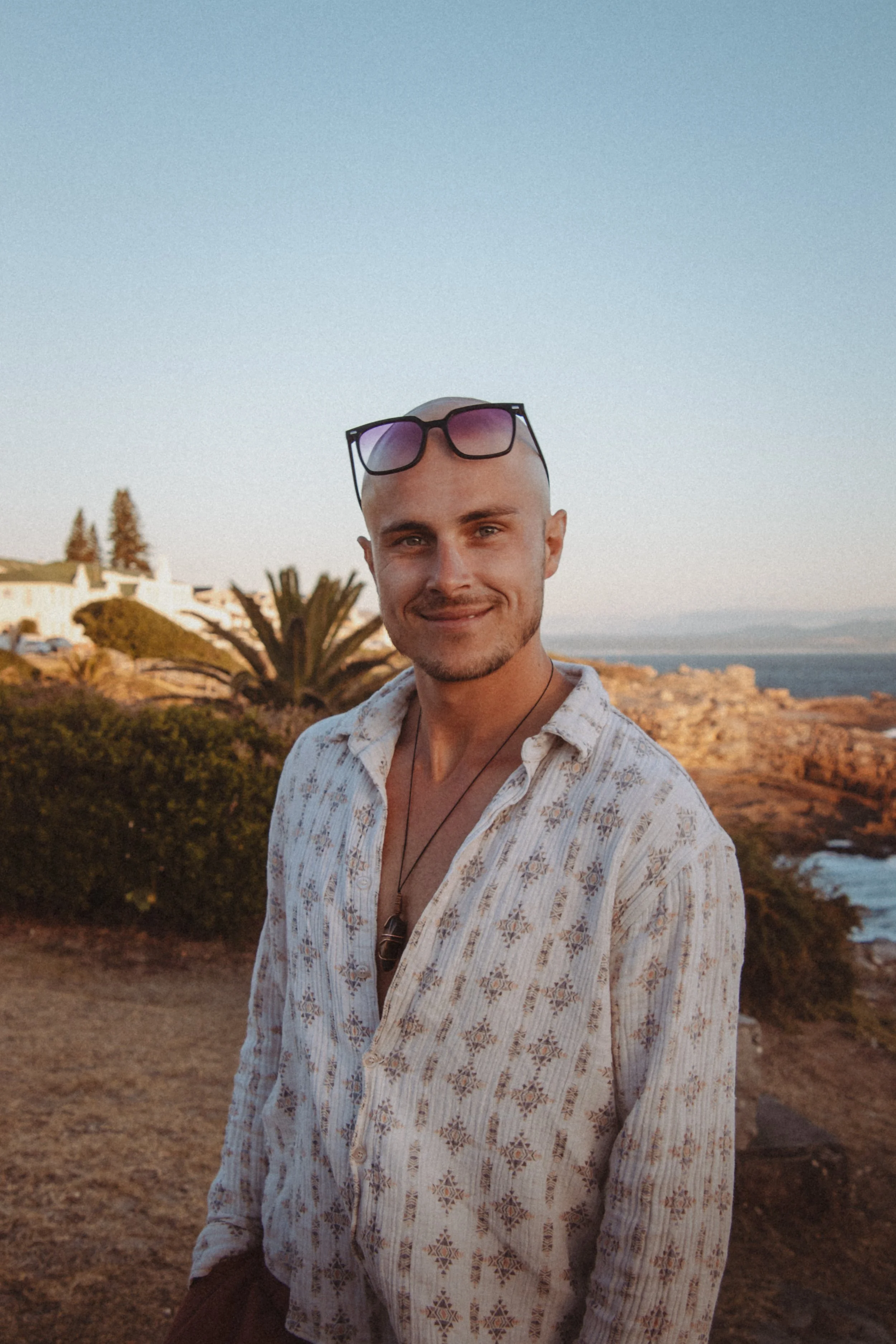 Ein junger Mann mit Sonnenbrille auf dem Kopf, lächelnd, vor einer Küstenlandschaft bei Sonnenuntergang.