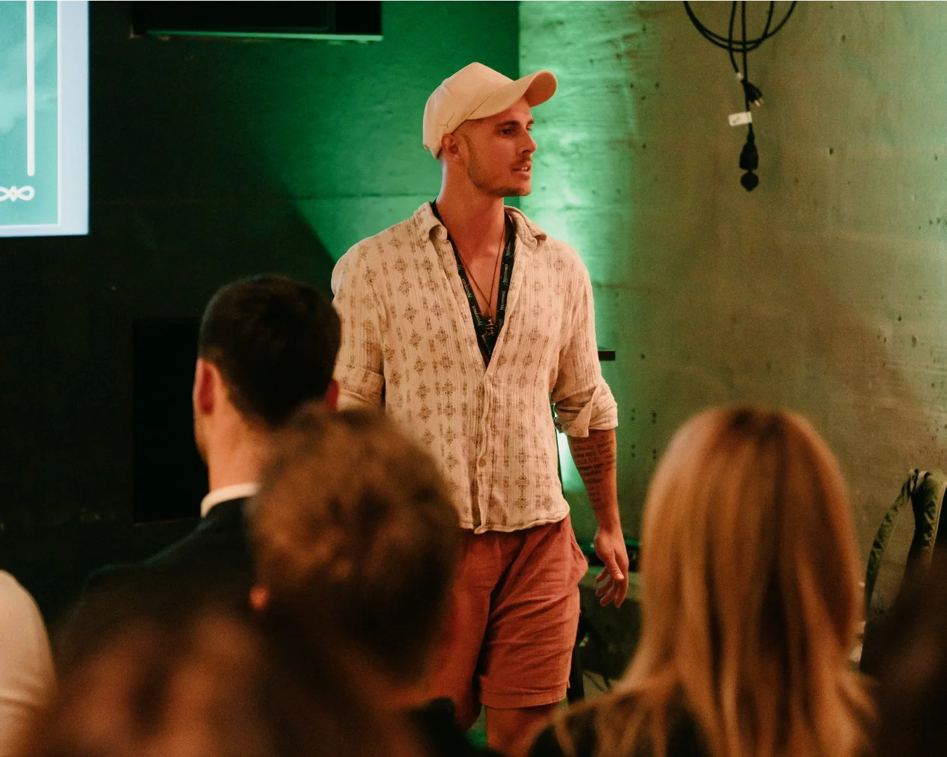 Ein junger Mann steht vor einer Gruppe und hält eine Präsentation. Er trägt eine Kappe, ein Hemd und Shorts. Die Szene ist in einem Raum mit grüner Beleuchtung.