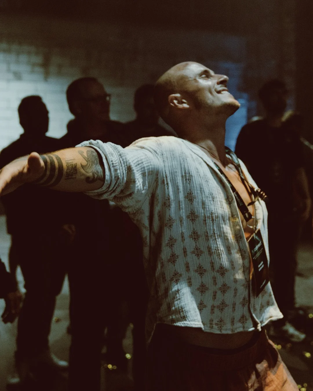 Ein Mann mit Tattoos auf dem Arm, lächelnd und mit offenen Armen, steht in einer Gruppe dunkler Silhouetten. Er trägt eine helle, gestreifte Jacke und eine Lanyard um den Hals, in einem dimmigen, urbanen Raum.