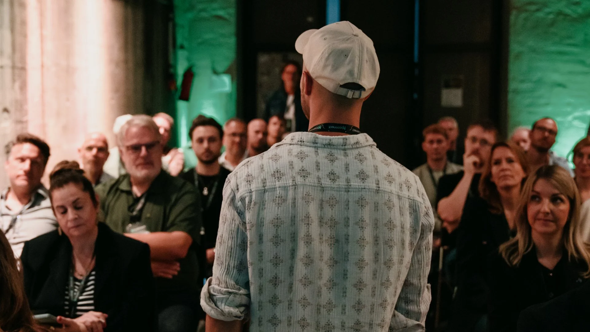 Mann trägt weißen Hut und Hemd mit Muster, spricht vor einer Gruppe von Zuhörern in einem dunklen Raum mit grünen Akzenten.