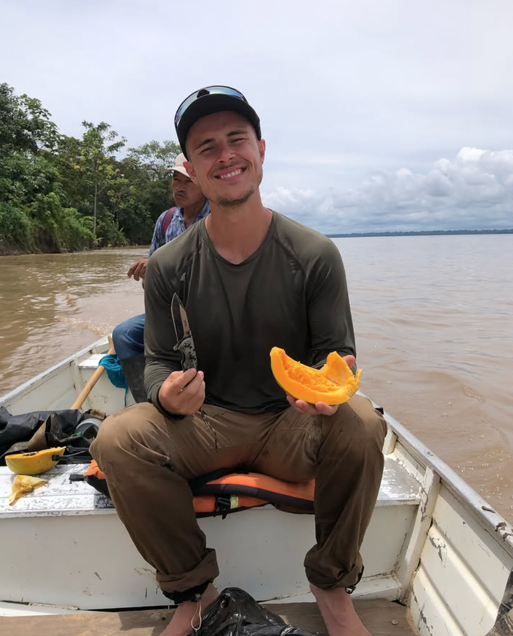 Mann sitzt in einem Boot auf einem Fluss und hält eine halbierte gelbe Paprika und ein Messer, umgeben von zwei weiteren Menschen bei bewölktem Himmel, mit grüner Natur am Ufer im Hintergrund.