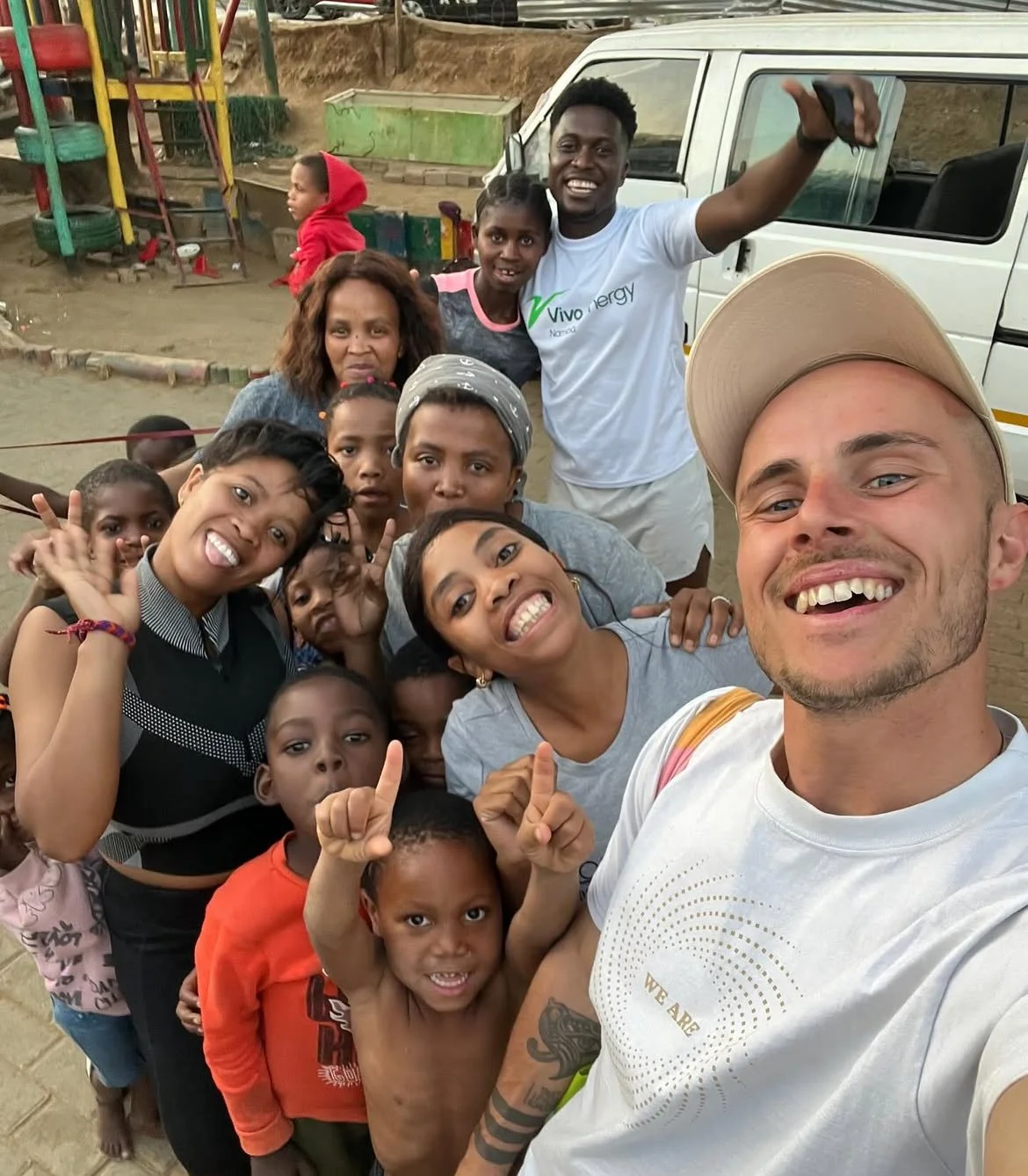 Gruppe von lachenden Menschen, Kinder und Erwachsene, die einen Selfie machen in einem offenen Raum mit Spielplatz im Hintergrund.