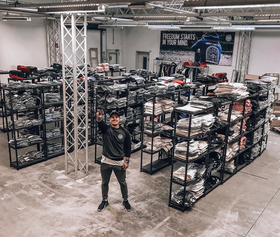 Mann in einem Lager mit mehreren schwarzen Regalen voller Kleidung, darunter Hemden und Hosen, und einem großen Banner an der Wand, das 'FREEDOM STARTS IN YOUR MIND' zeigt.