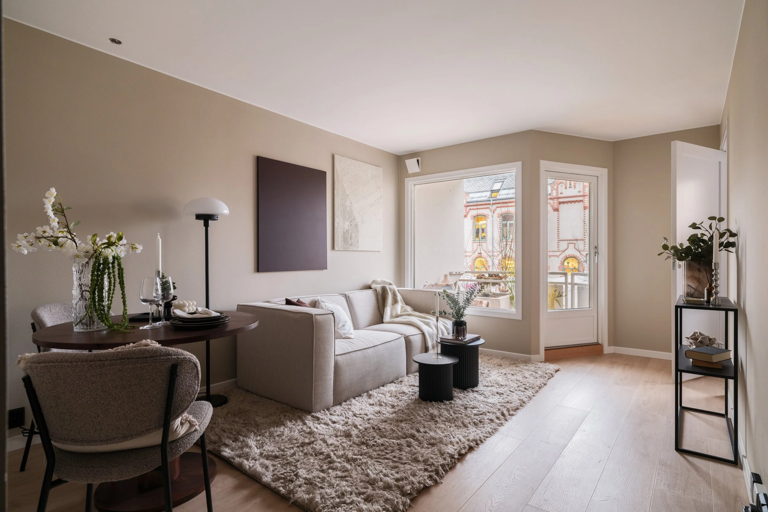 Stue med lys beige vegger, stor sofa, rundt bord med blomster, kunst på veggene, vindu og dør til balkong, tregulv og teppe, pyntede stoler og dekorasjon.