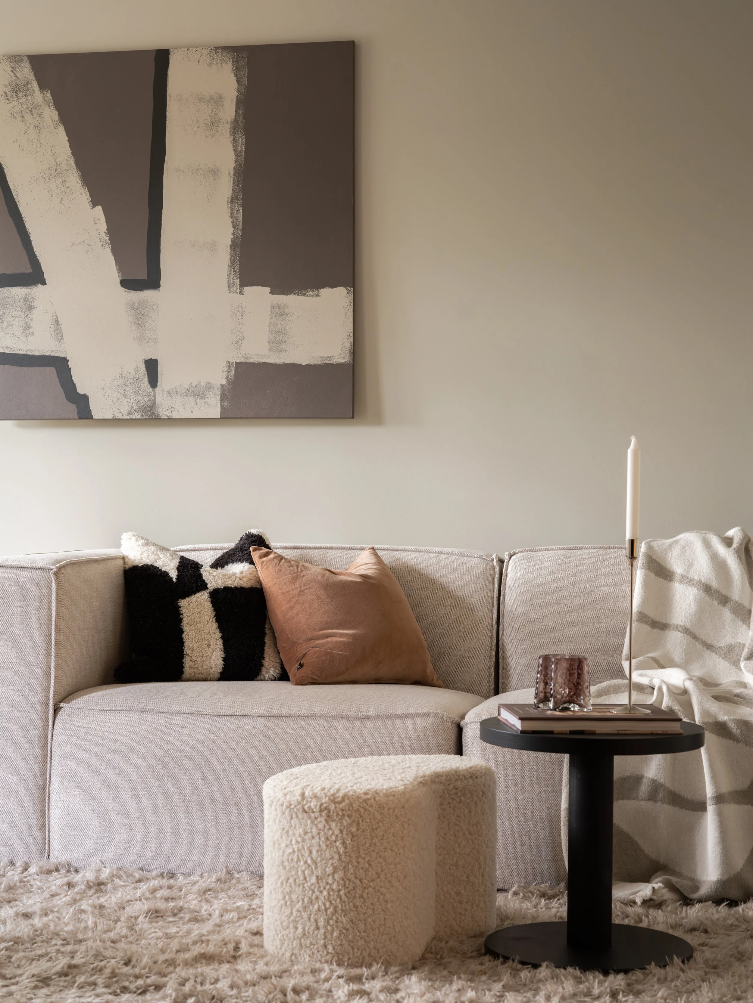 Et stueområde med en beige sofa, pyntet med to puter, en i svart og kremfarget mønster og en i lys rosa. En svart sidebord med bøker og lysestake, og en liten rund, kremfarget puff foran. Veggen har et abstrakt kunstverk i grå og hvite toner, og en lang hvit lysestake med ett hvitt lys står på bordet. Det er en myk, lys brunt teppe på gulvet.