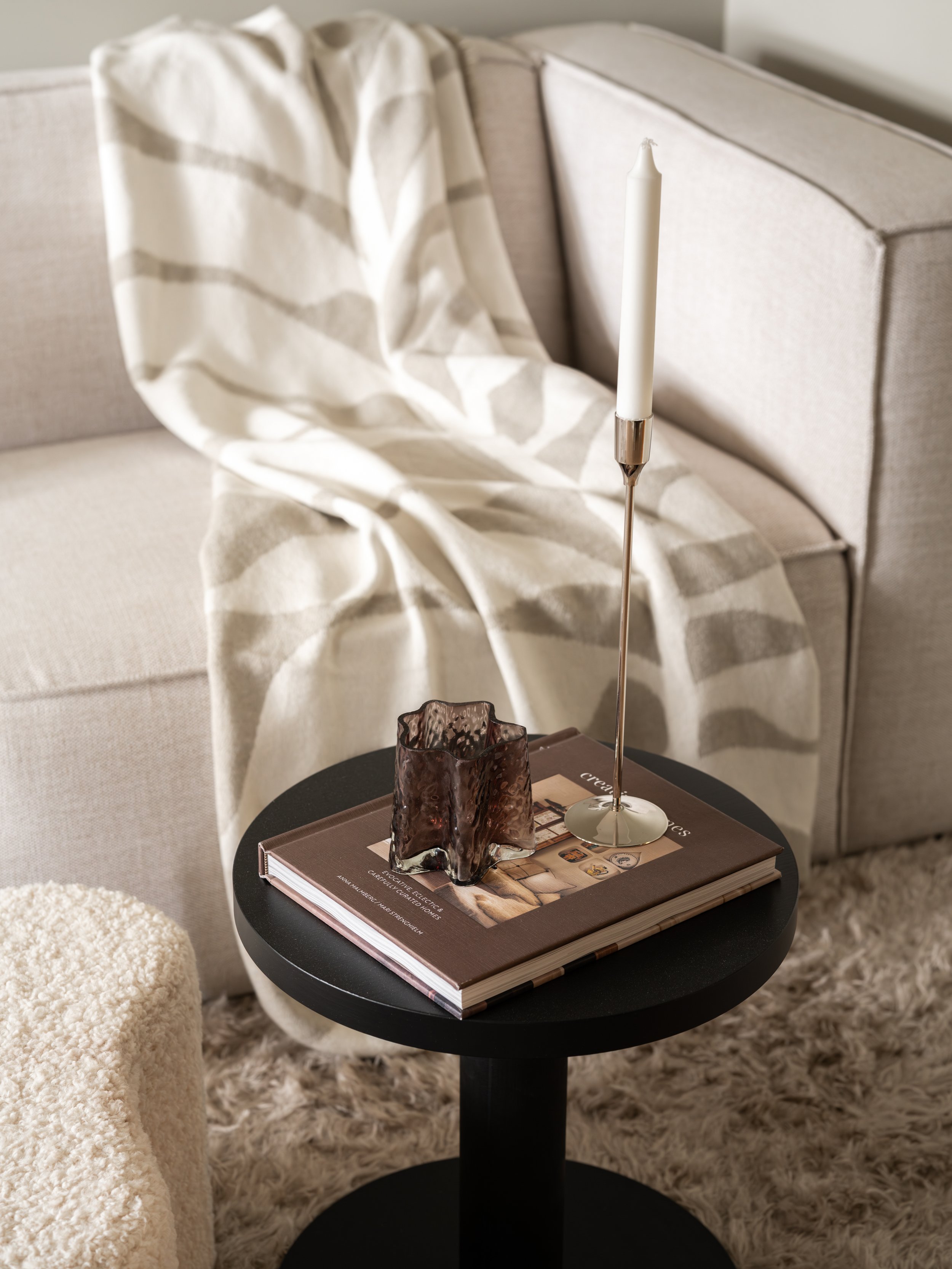 Et svart bord med en bok, en glasslampe og et lysestake står foran en beige sofa med en hvit og grå pledd.