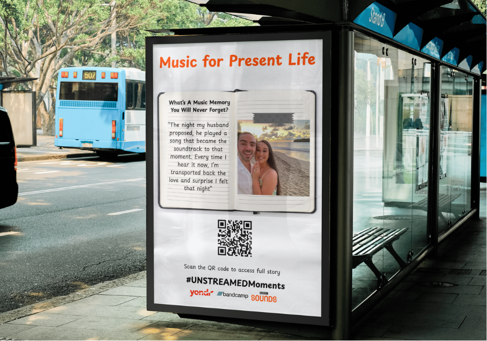 Advertisement on a bus shelter advertising music memories with a photo of a smiling couple on a beach at sunset, a QR code, and the hashtag #UNSTREaMEDMoments.