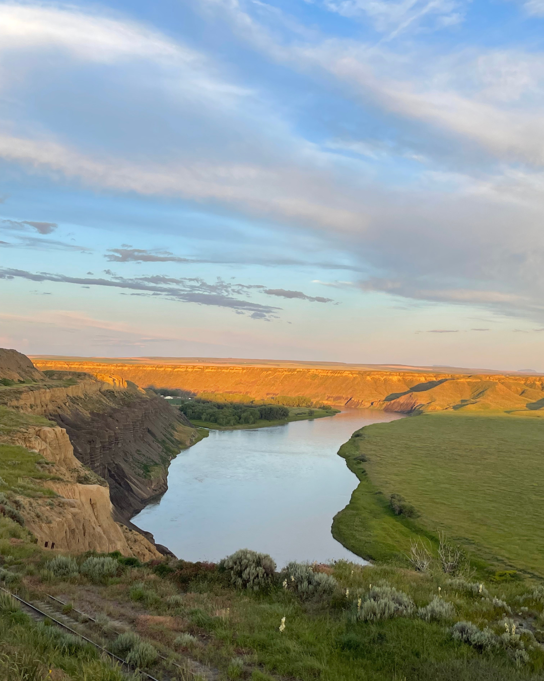 A wide river flowing through a lush green valley with steep rocky cliffs on the left and rolling hills on the right, under a sky with scattered clouds during the daytime.
