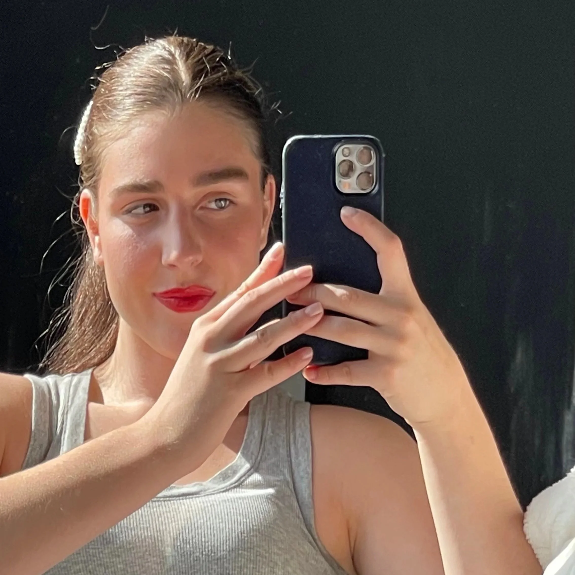Close-up selfie of a young woman with long brown hair, wearing a light-colored tank top, red lipstick, and holding a smartphone in front of a mirror.
