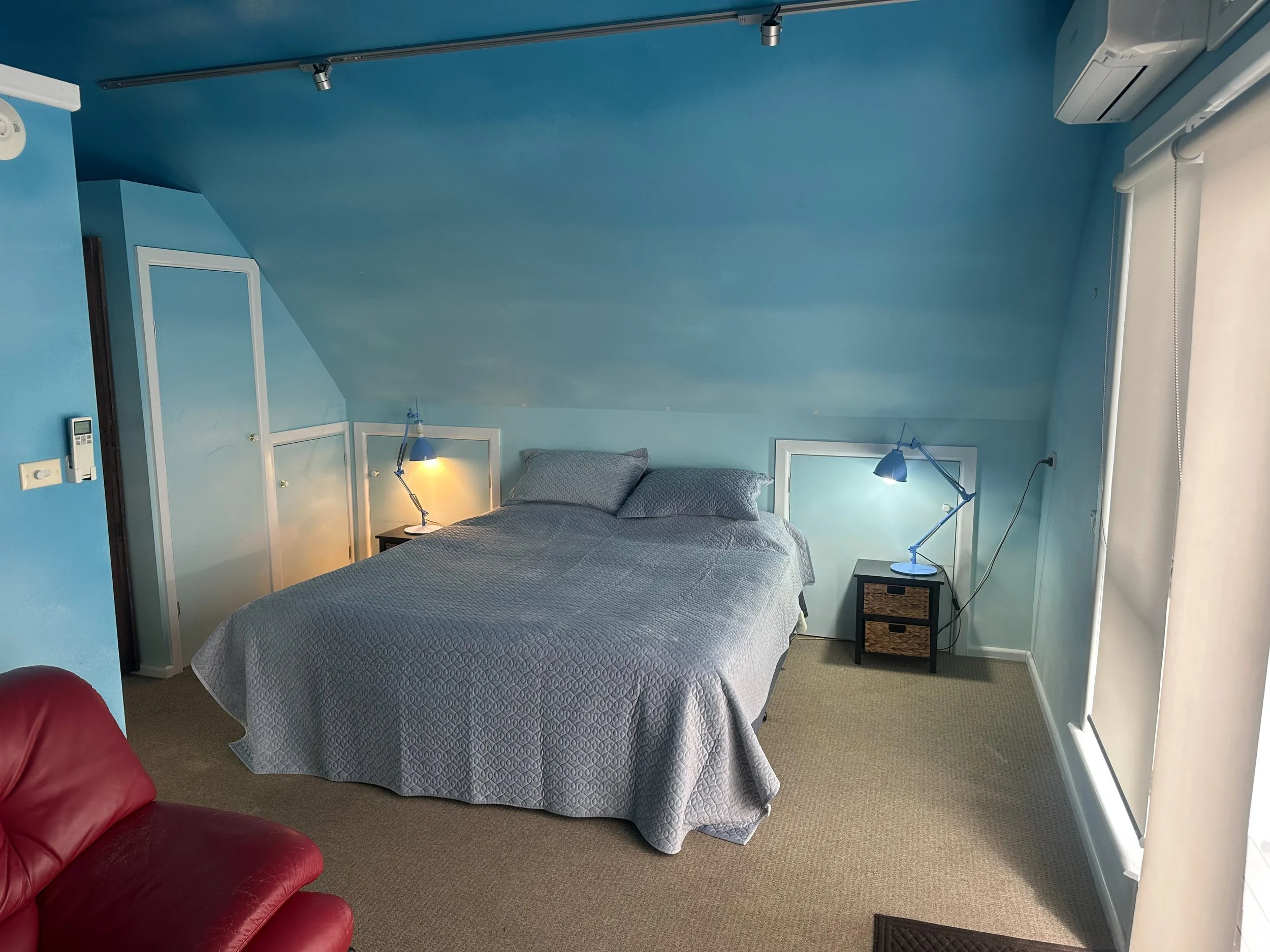 A cozy bedroom with a bed in the center, two nightstands with blue reading lamps, an air conditioning unit, a red armchair, and a large window covered with a white curtain.