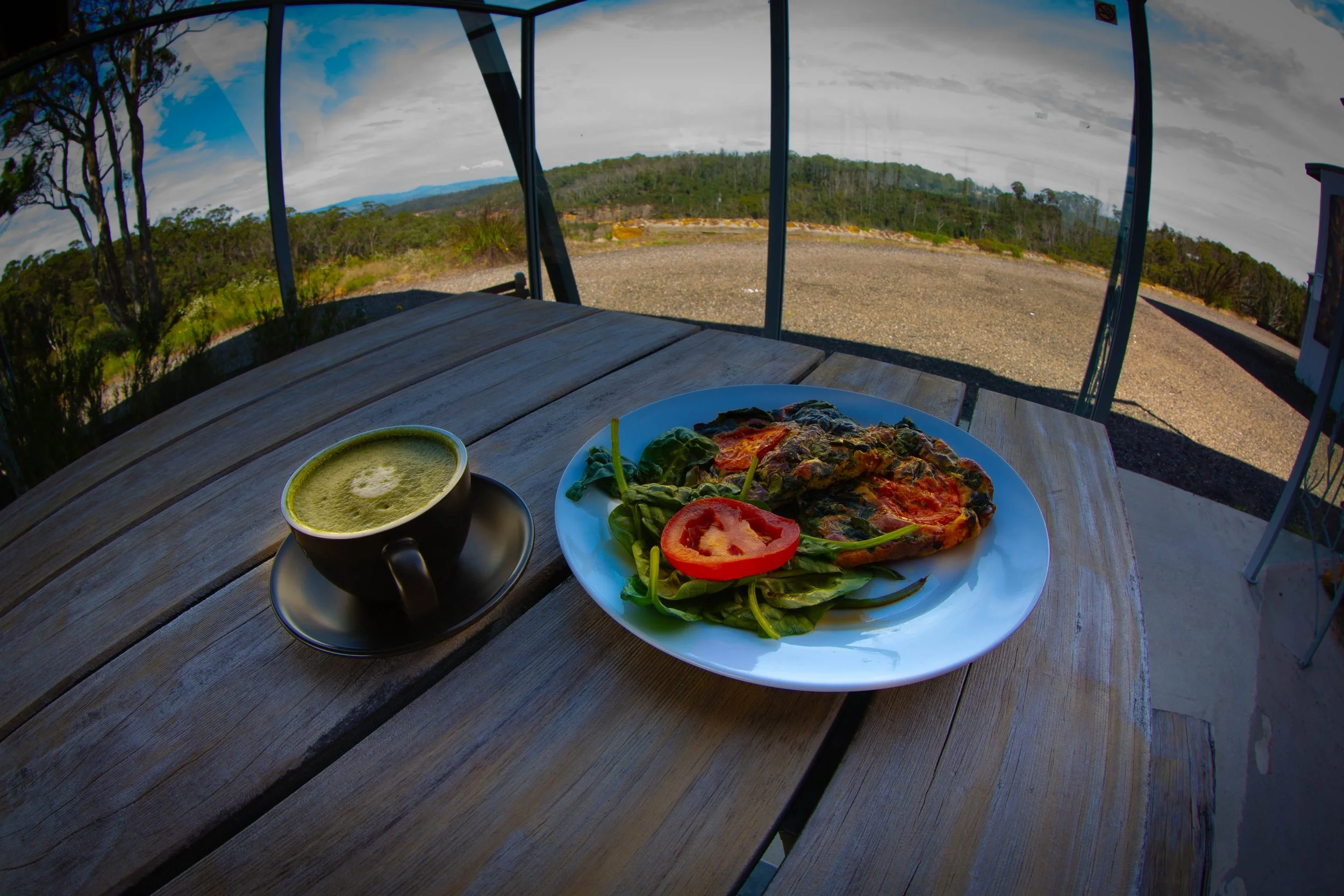 A meal on a wooden table includes a slice of vegetable quiche, a small salad with greens and a tomato slice, and a cup of green tea, with a landscape view of trees and a cloudy sky outside.