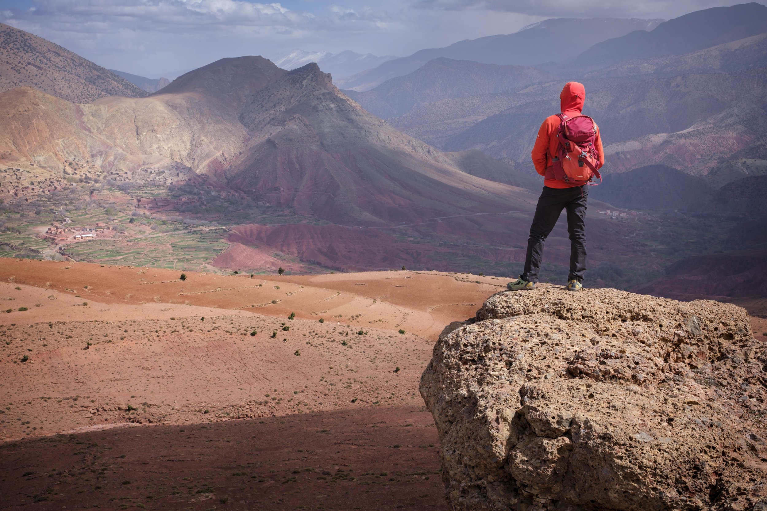 Person in red jacket and backpack standing on a large rock, overlooking a valley with mountains in the background.