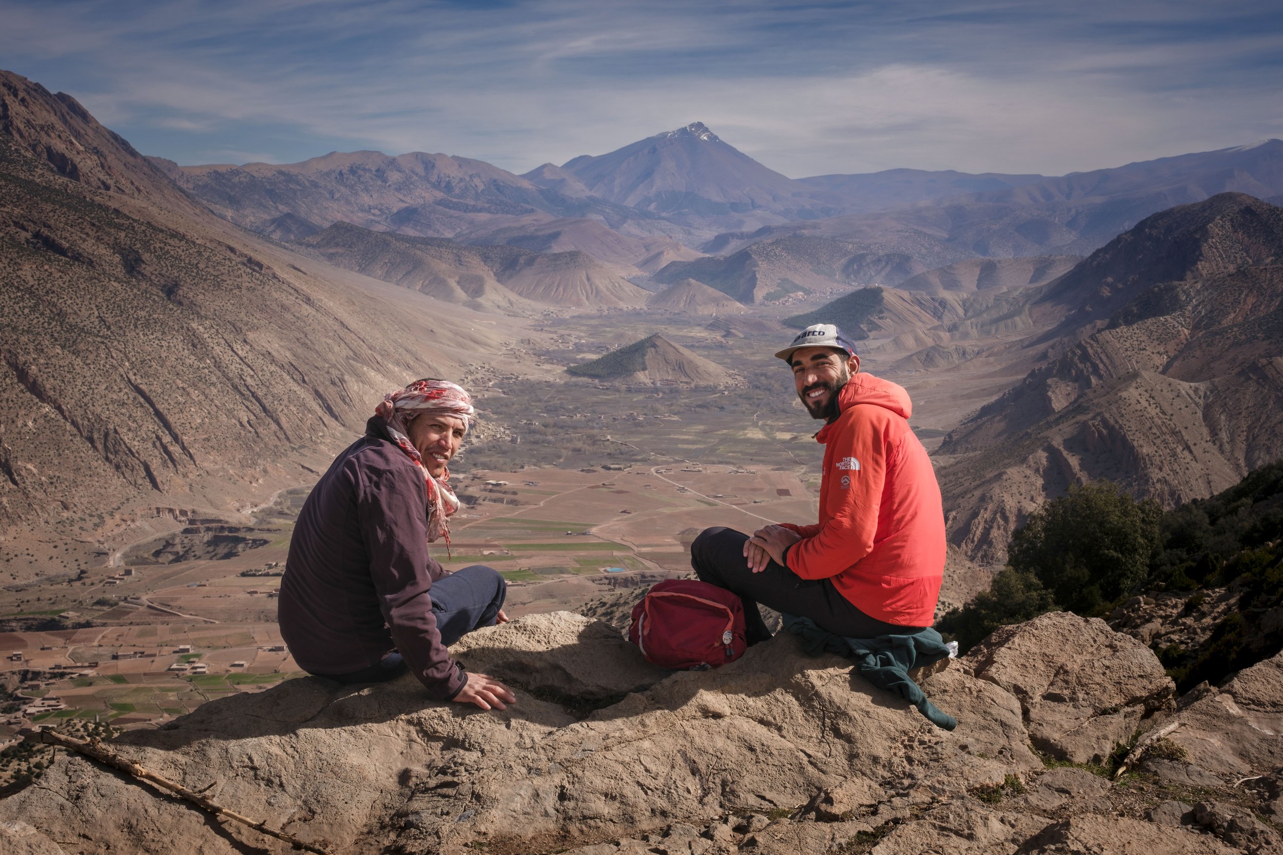 Two men resting on a rocky ridge overlooking the wide Aït Bouguemez Valley in Morocco’s High Atlas Mountains.