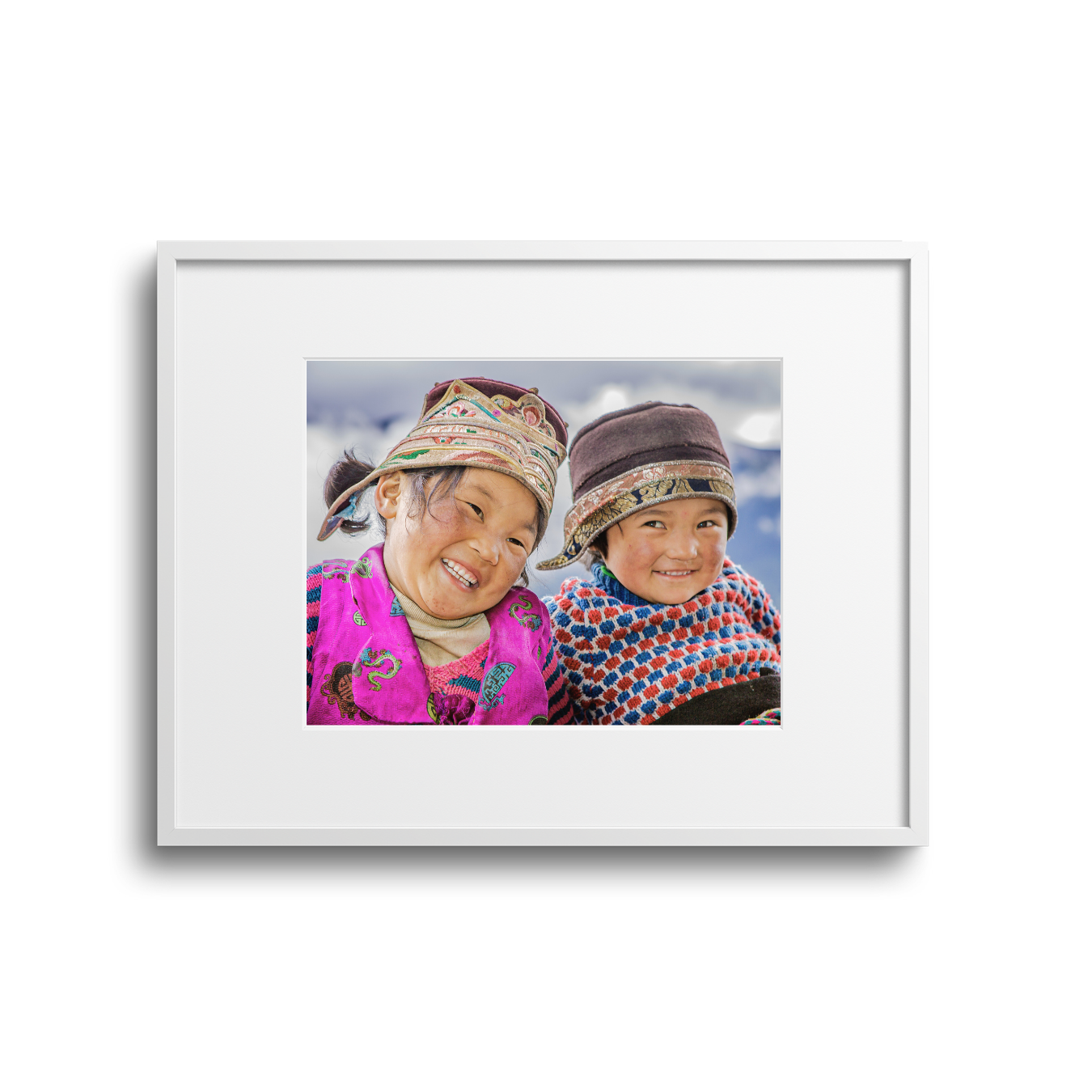 Two smiling children wearing traditional hats and colorful clothing, outdoors with mountains and cloudy sky in the background.