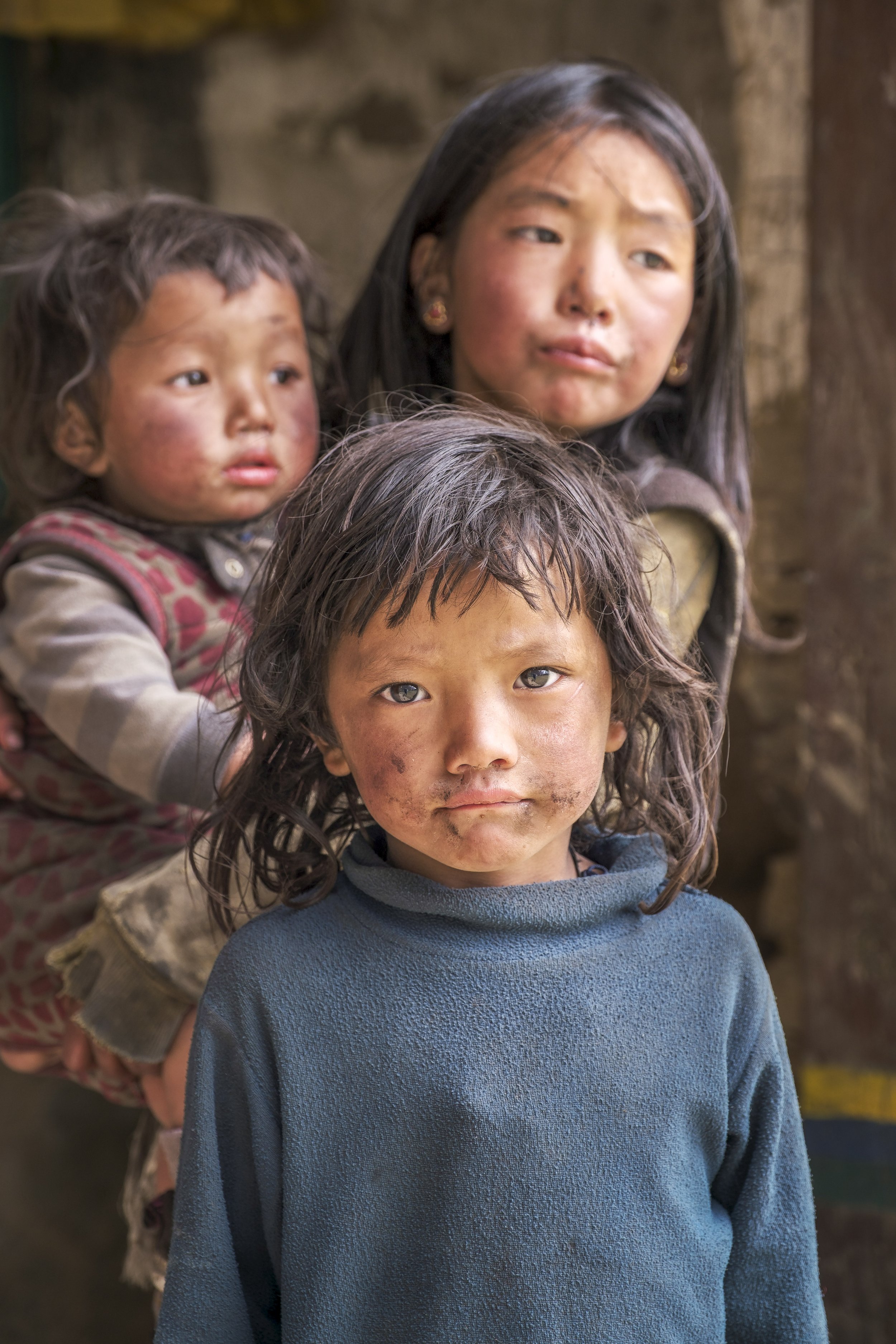 Three siblings stand outside the old monastery in Pangdun. The eldest girl watches over the younger ones as she does most days, where childhood often begins with responsibility in the valley. Tsum Valley, Nepal. Photo By Steve Fagan.