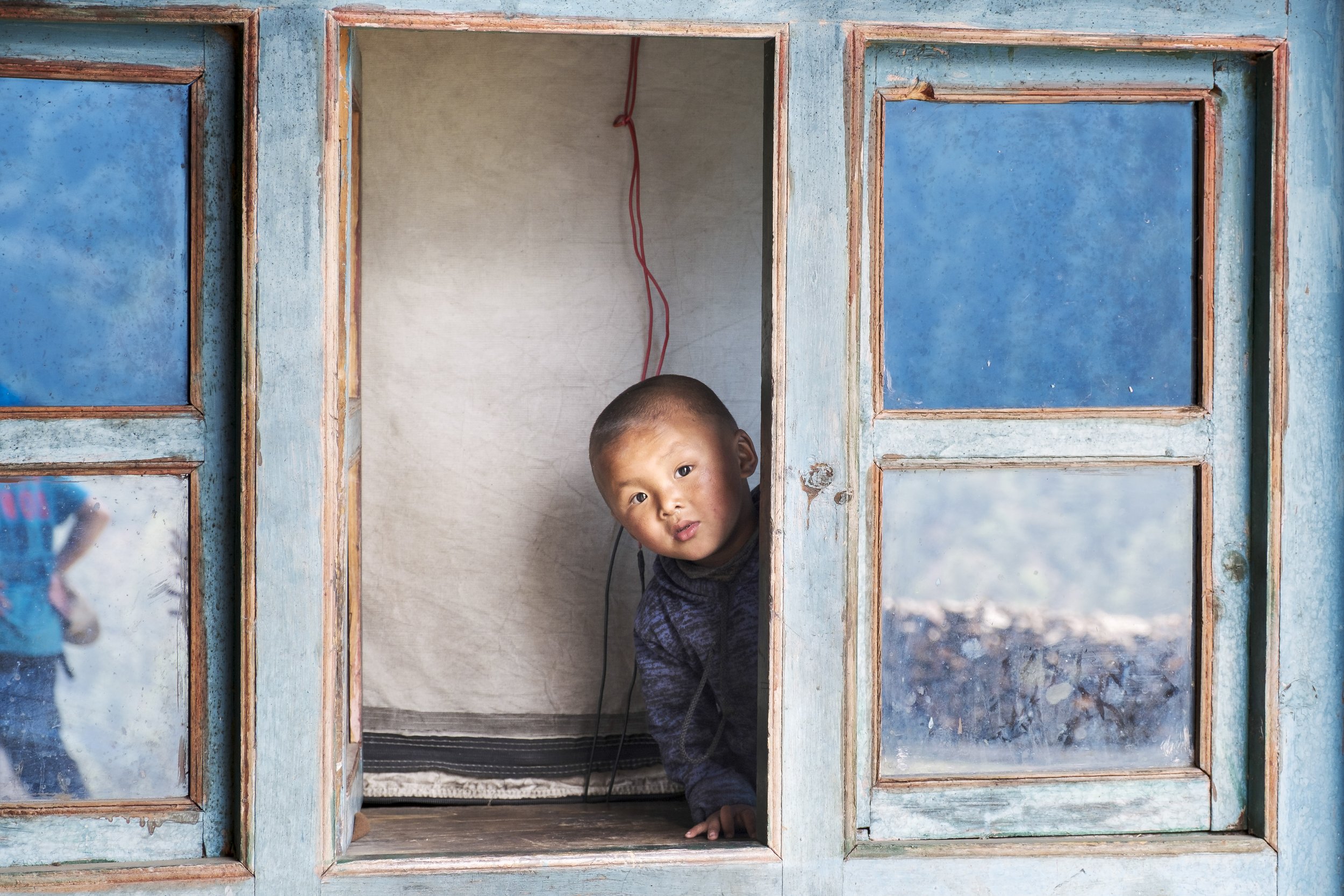 Morning light falls across the weathered blue window of Tashi Delak Guesthouse. The owner’s grandson leans into the frame during his playtime, watching the courtyard where trekkers come and go. Tsum Valley, Nepal. Photo By Steve Fagan.