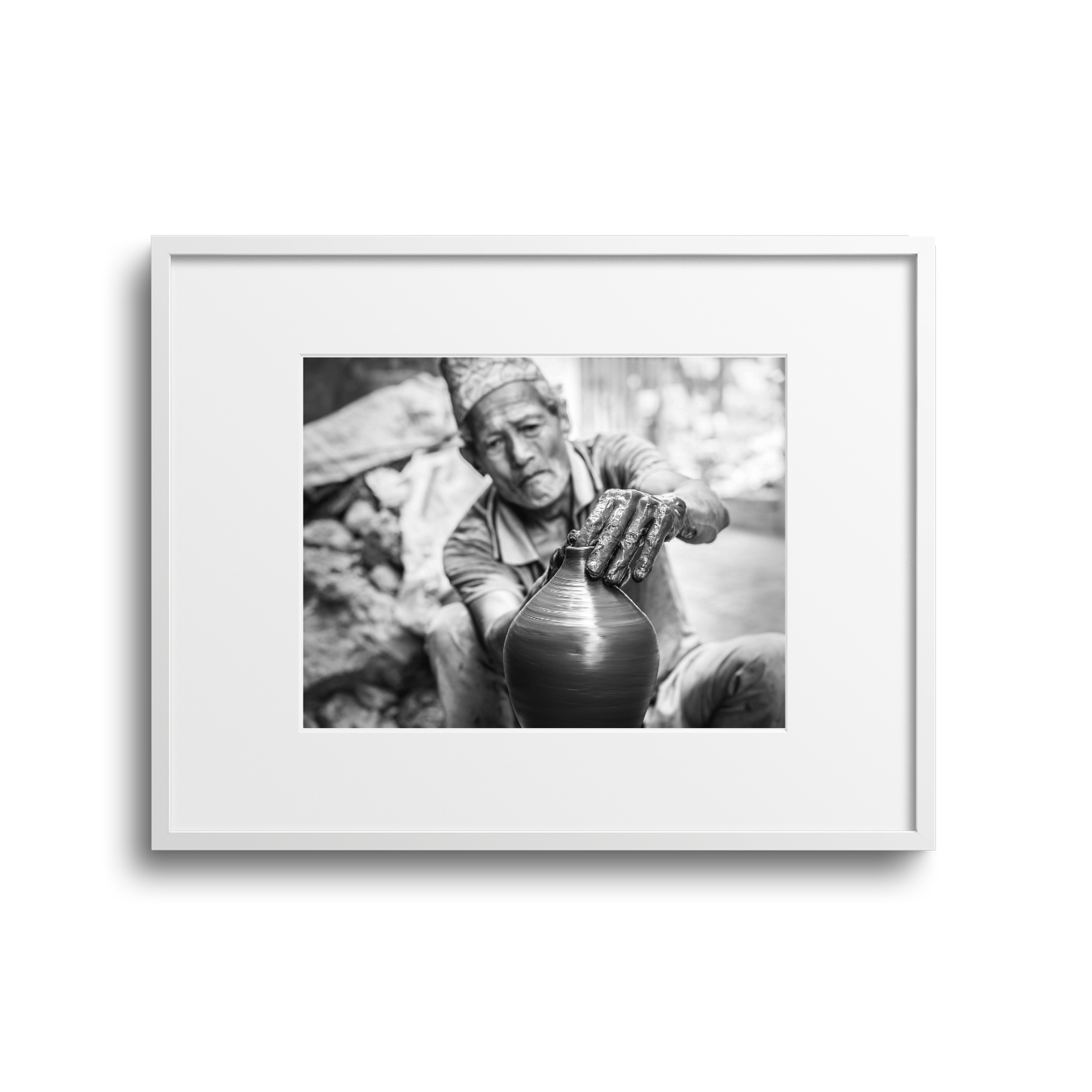 A black and white photo of an elderly man shaping a clay pot with his hands, wearing a traditional hat and sitting outdoors.