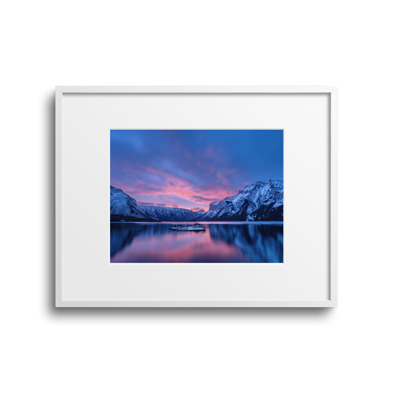 A framed landscape photo of a mountain lake during sunset or sunrise, with snow-capped mountains on either side and a colorful sky reflected in the water.