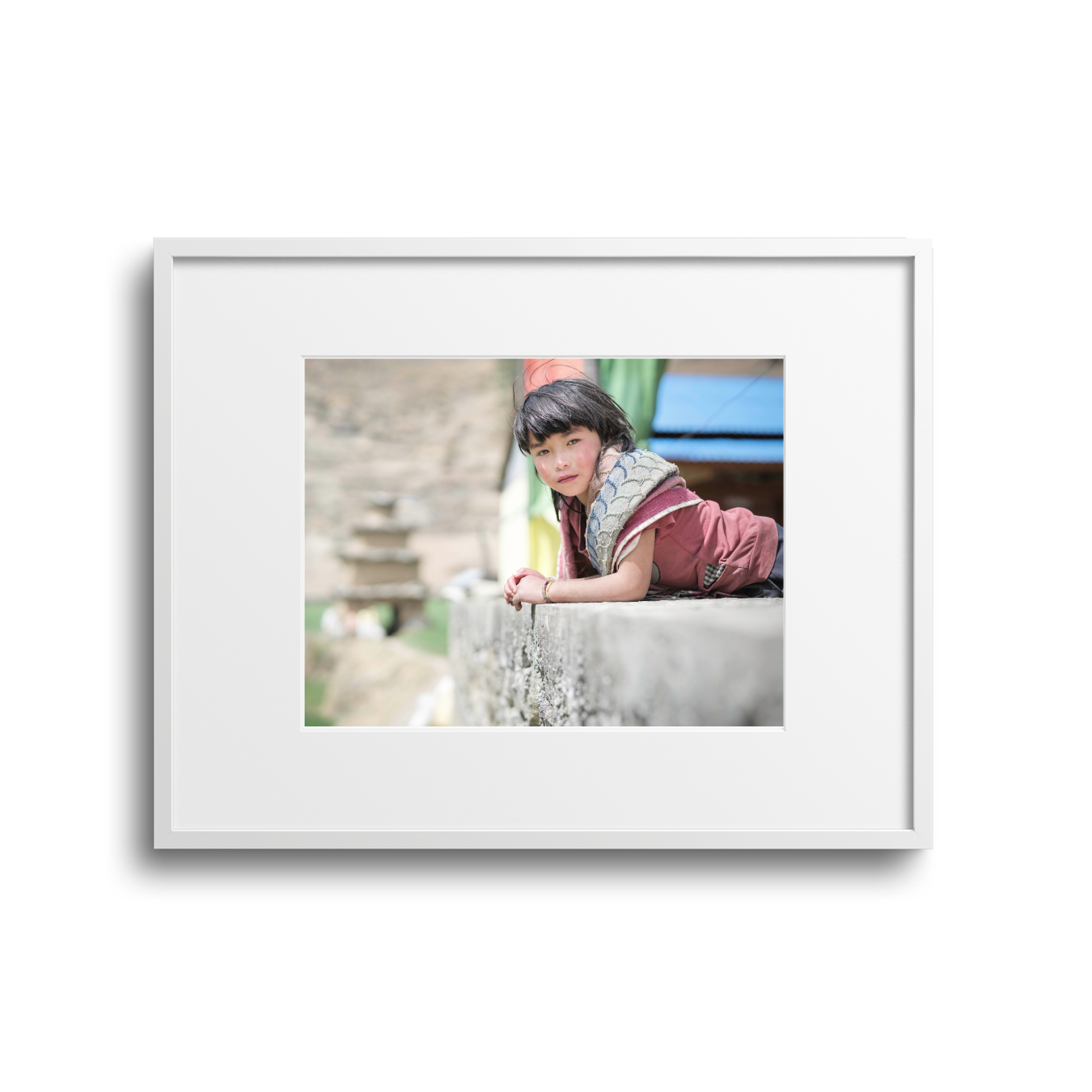 Young girl with dark hair and bangs lying on a stone ledge outdoors, wearing a maroon shirt and a grey scarf, looking at the camera.