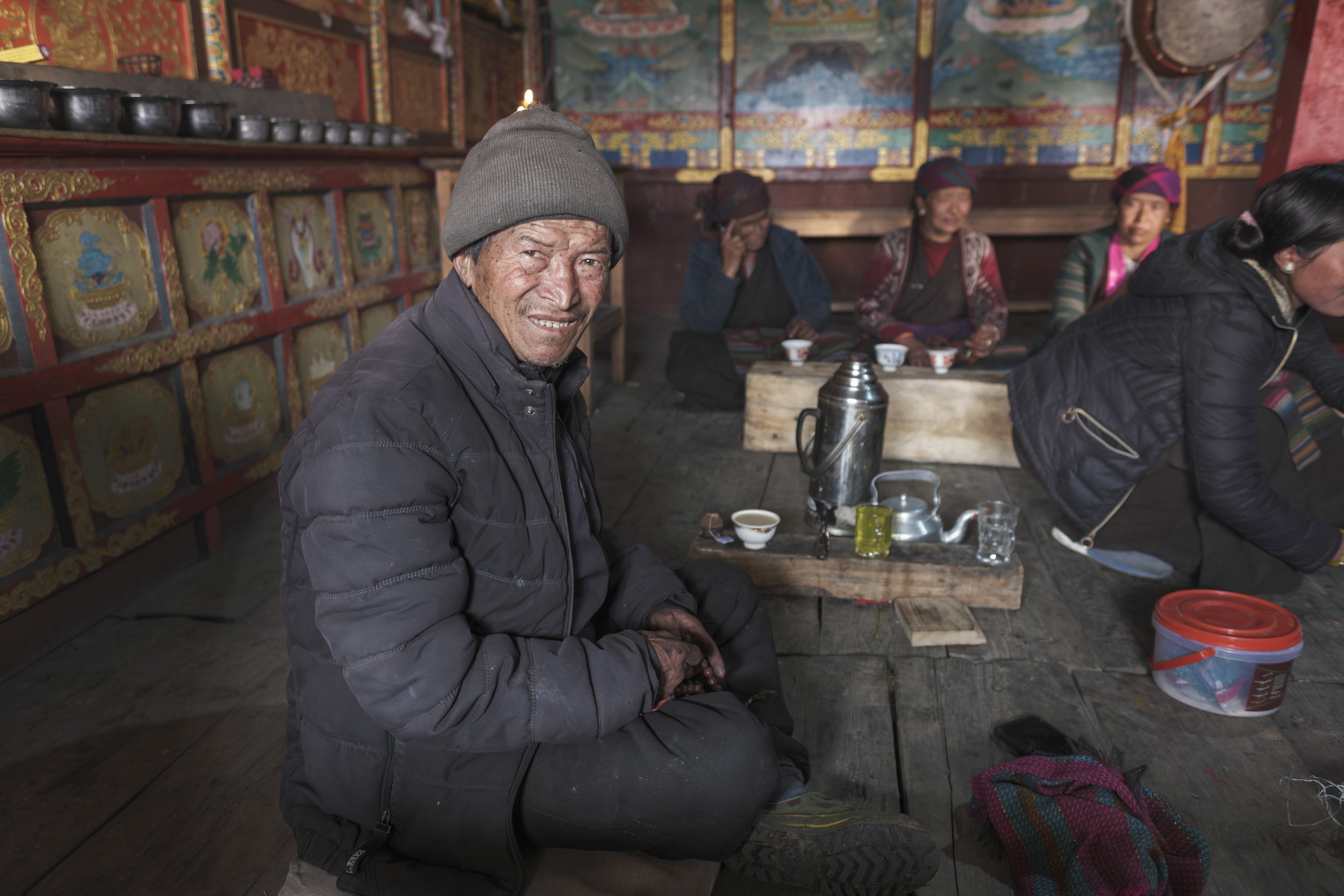 Man seated inside a monastery beside a kettle and cups used for communal tea in Tsum Valley, Nepal.