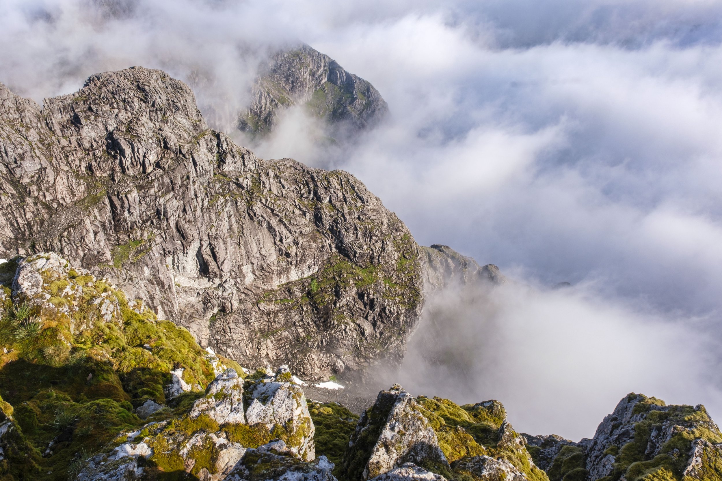 Mountain peaks covered in fog with rocky terrain and green moss in the foreground.