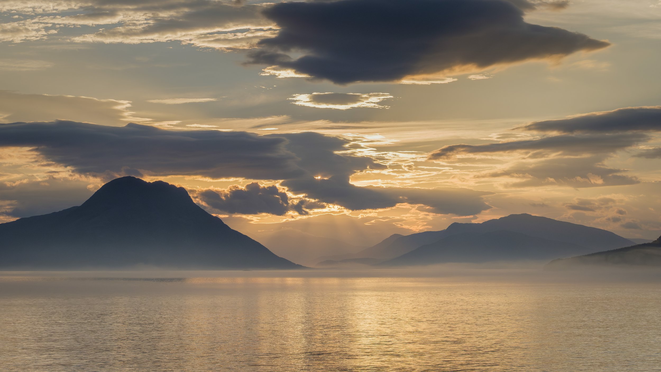 Sunset over a mountain landscape with a body of water in the foreground, partially covered by mist and clouds in the sky.