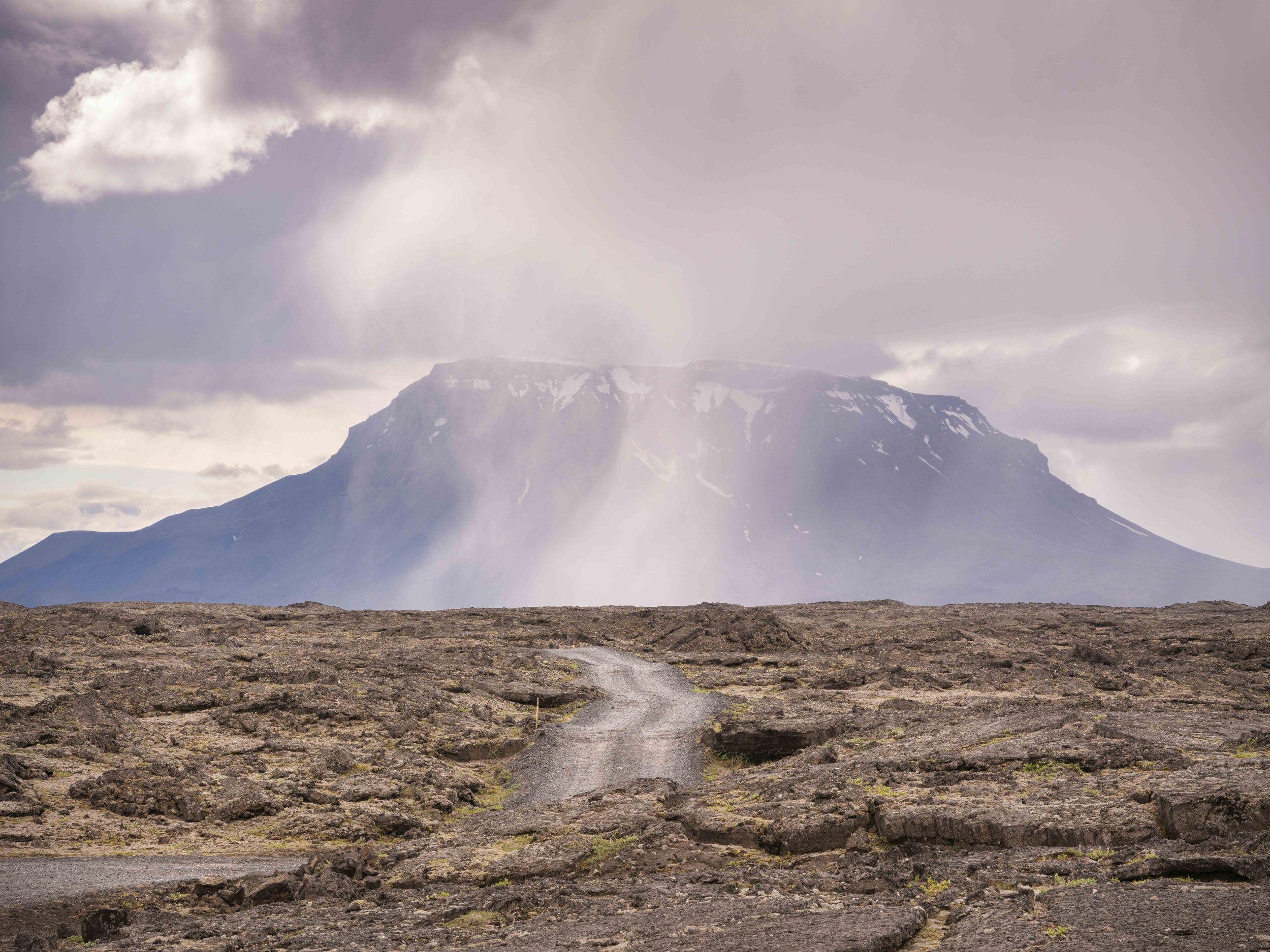 A rocky dirt pathway leads toward a mountain in the distance with patches of snow, under a cloudy sky with sunlight breaking through.
