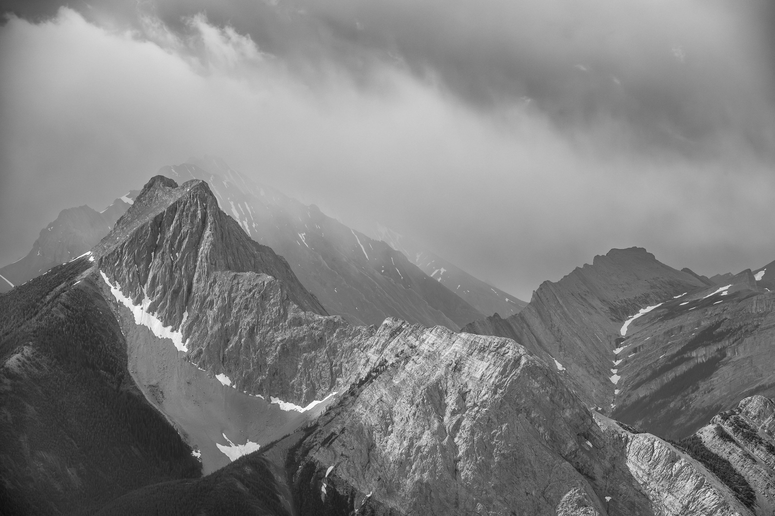 Black and white photograph of rugged mountain peaks under a cloudy sky with some snow patches on the slopes.