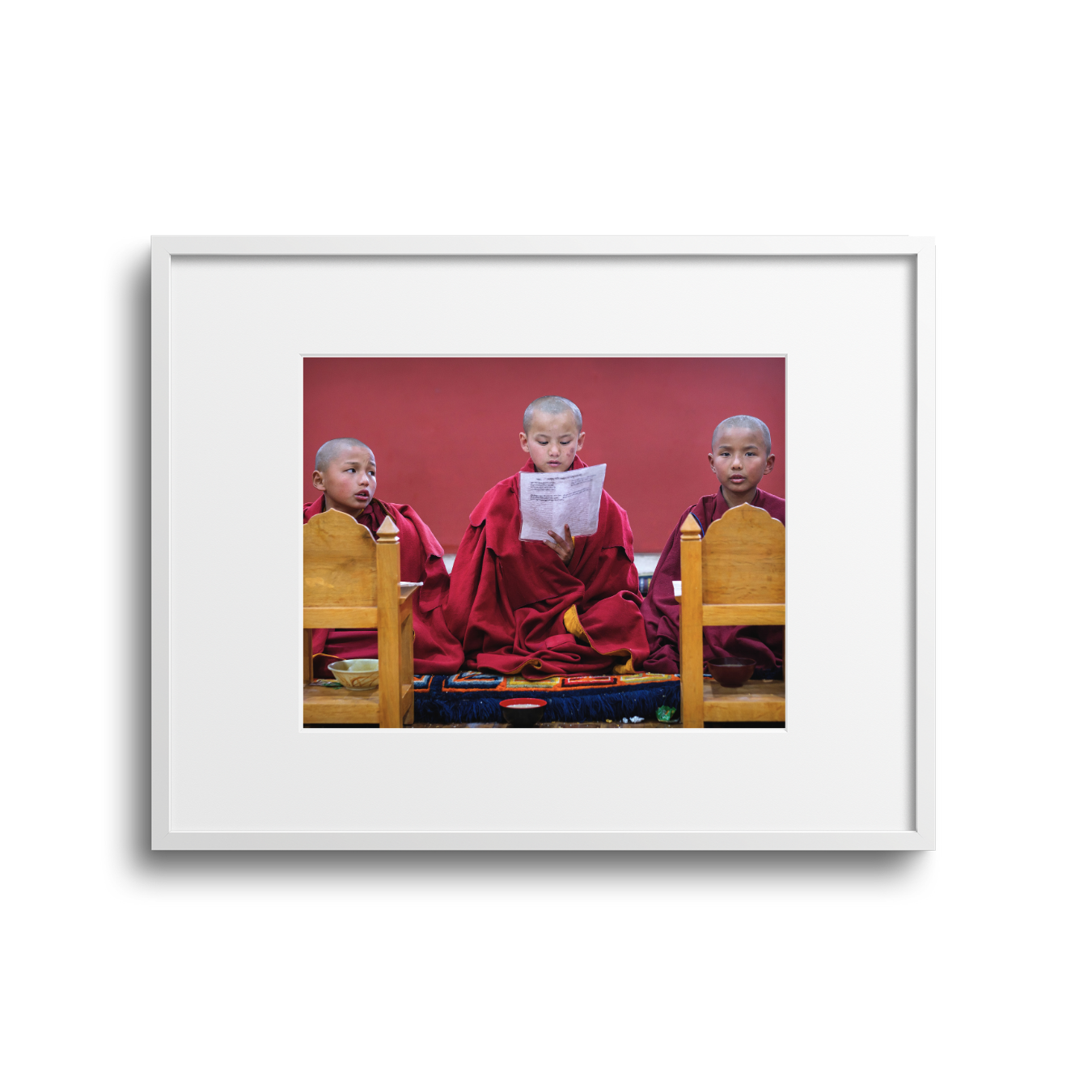 Three young monks in maroon robes sitting on the floor with bowls in front of them, one of them reading a paper, against a red background.