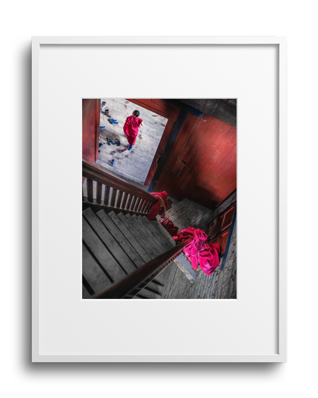 A person dressed in a red garment walking on a wooden floor at the bottom of a staircase, with shoes scattered around. The photo is taken from the top of the staircase looking downward, with parts of the staircase visible.