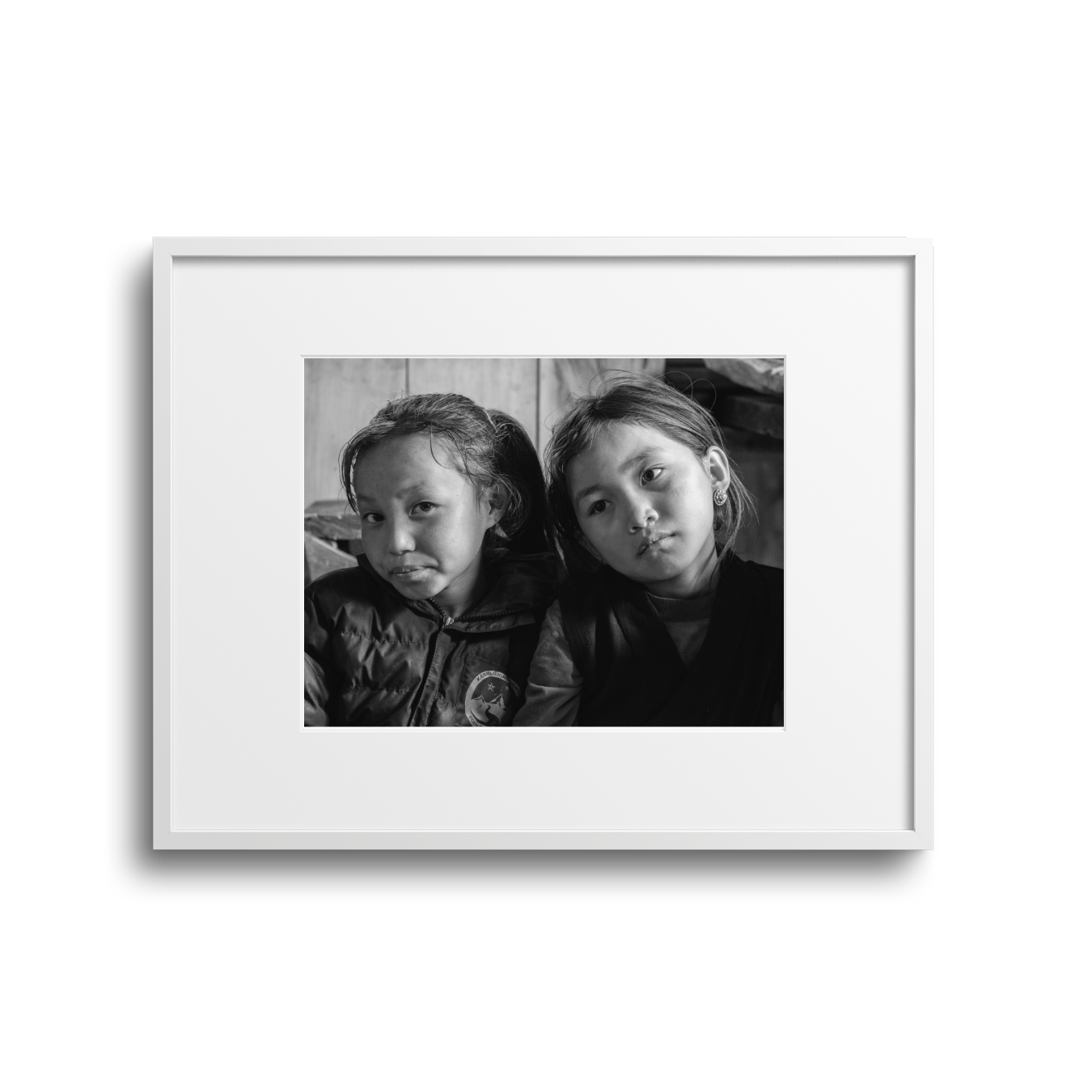 Black and white photo of two young girls sitting close together, one with her tongue out, inside a white picture frame.