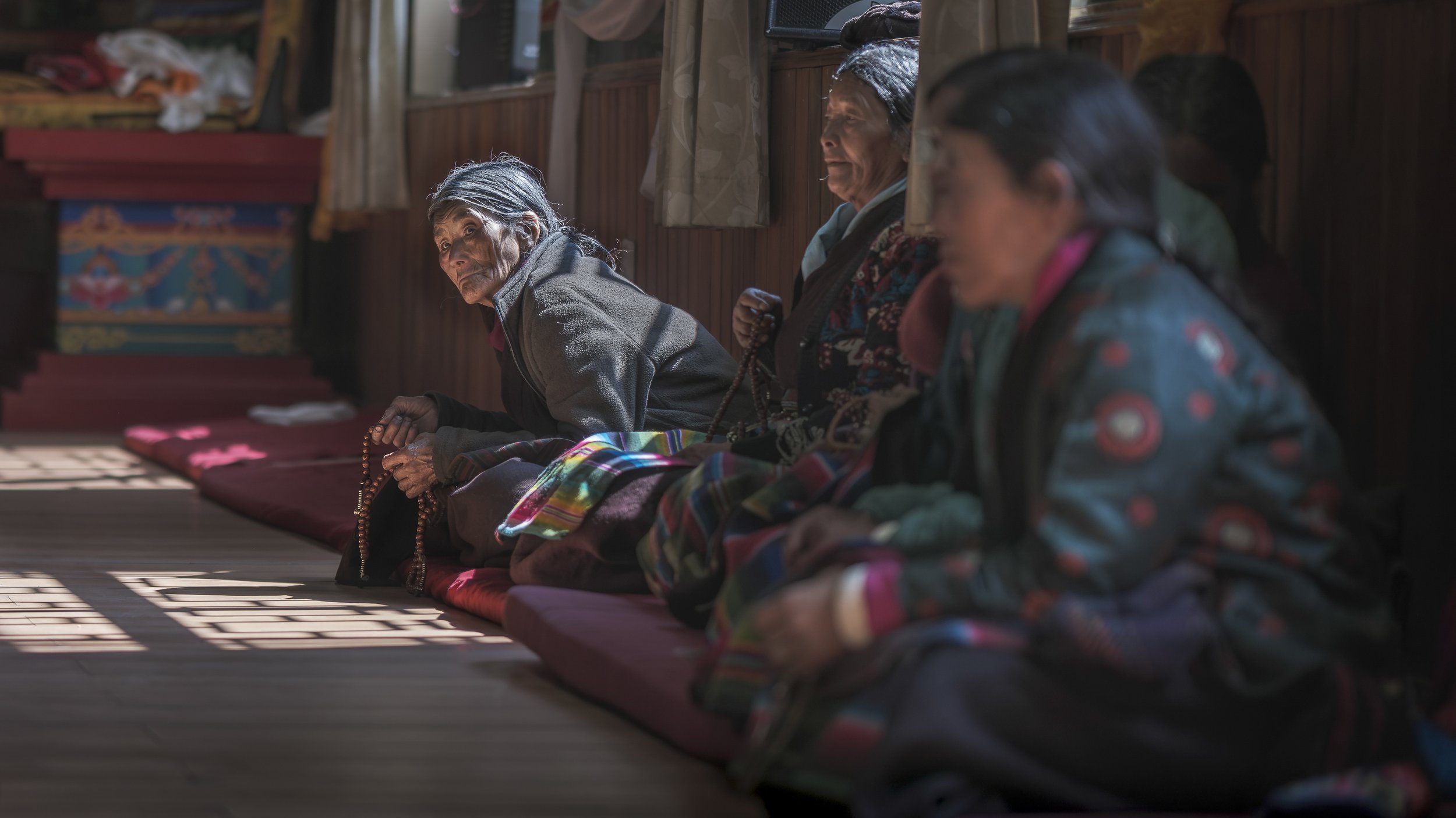An elderly woman sits quietly in prayer at Rachen Nunnery during the first death anniversary puja of His Holiness Zopa Rinpoche. Prayer beads rest in her hands as light falls across the wooden floor. Devotees have walked from Chekampar to Nile to be 