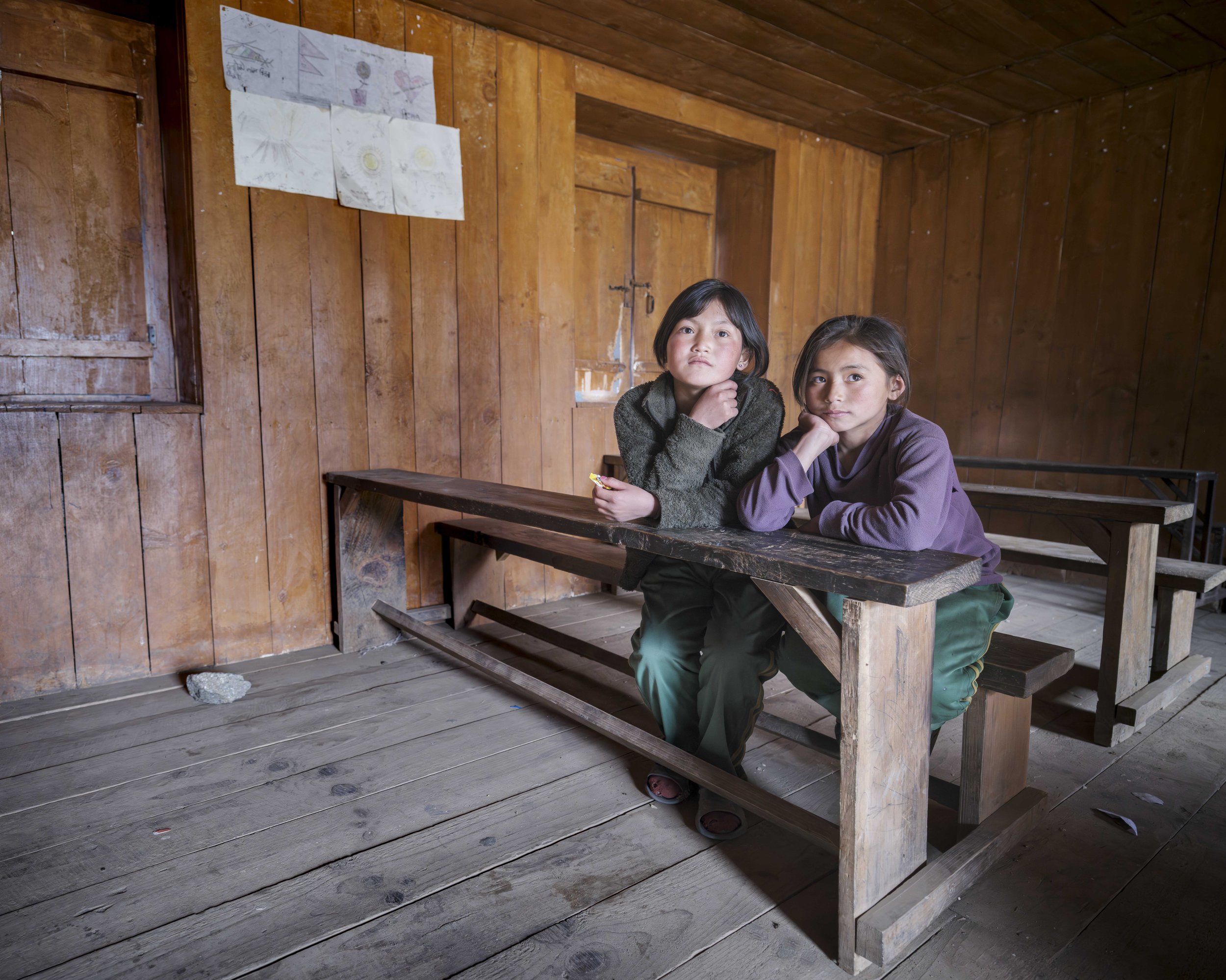 Two young girls sit inside a wooden classroom at Chumling School, where many students live as boarders during term. Between lessons, the room shifts from study to play. Tsum Valley, Nepal. Photo By Steve Fagan.