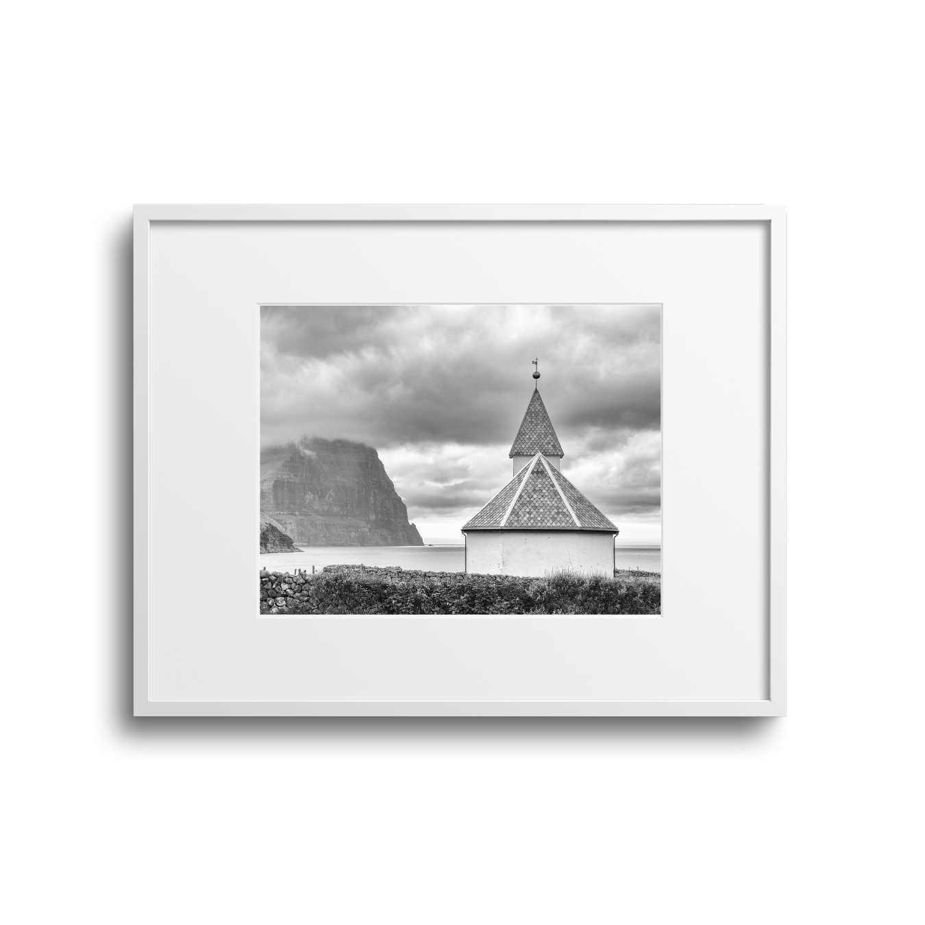 Black and white photo of a small church with a pointed steeple near the ocean, with cliffs in the background and cloudy sky.