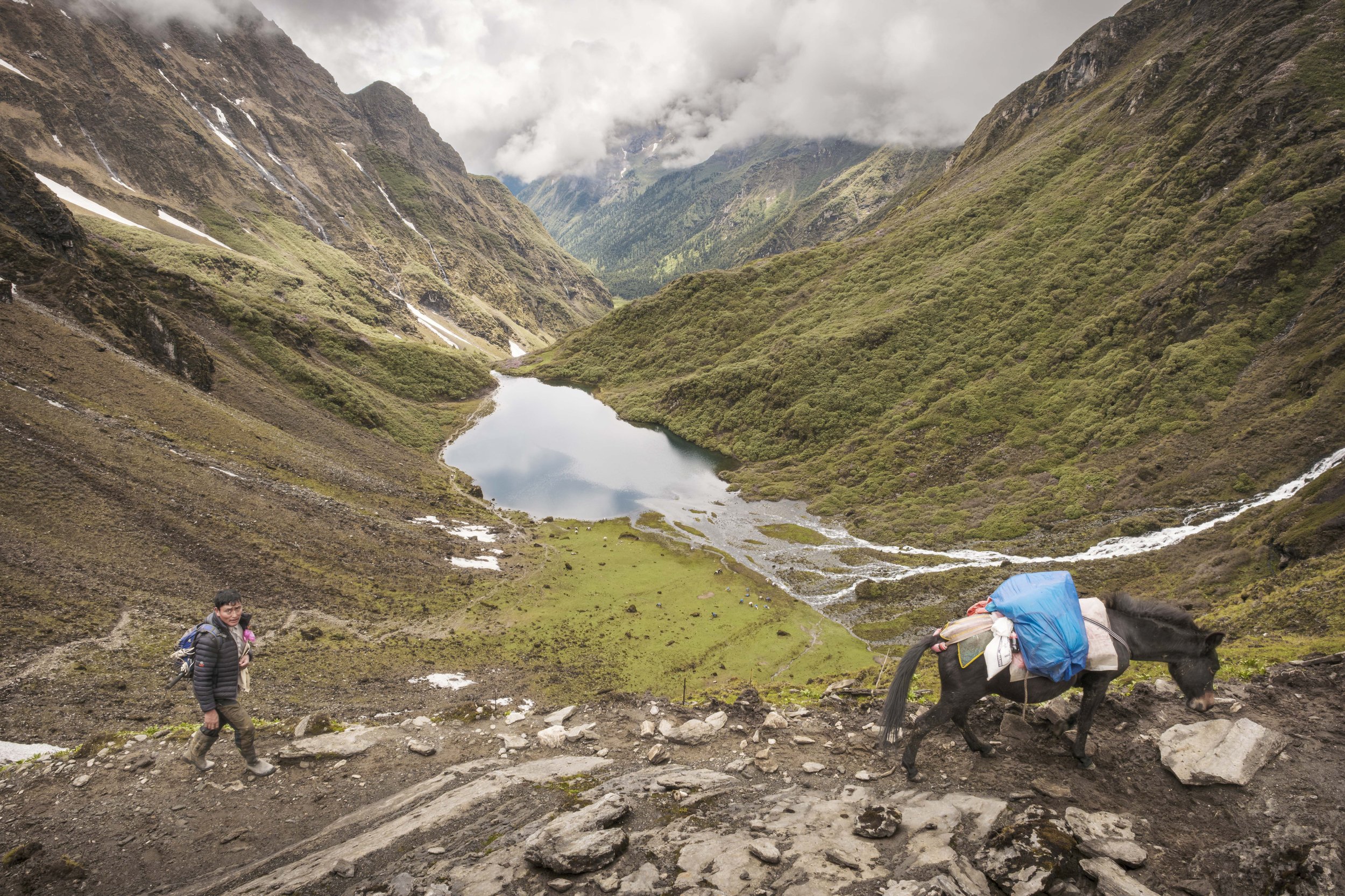 A hiker walking along a rocky trail beside a pack animal carrying supplies in a mountainous landscape with a lake and cloudy sky.