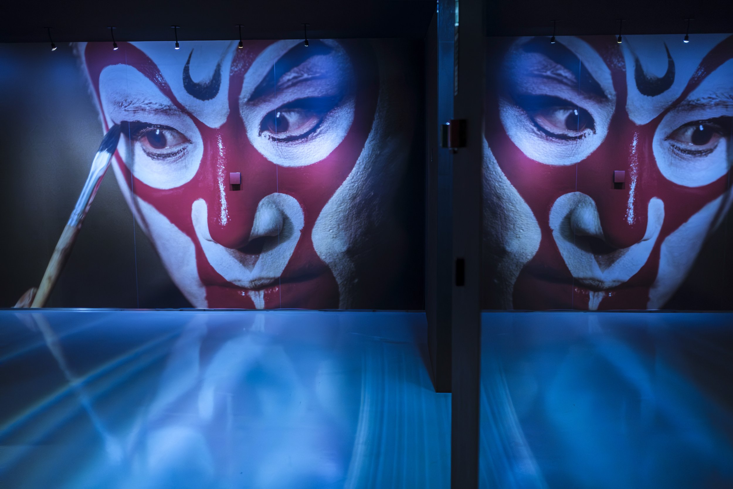 Large projected face with painted features reflected across mirrored walls in a dark exhibition space.