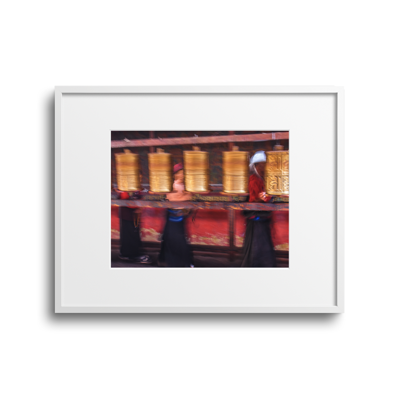 People walking past a wall of large, vertical prayer wheels at a Buddhist temple, with one person in a red robe and white hat.