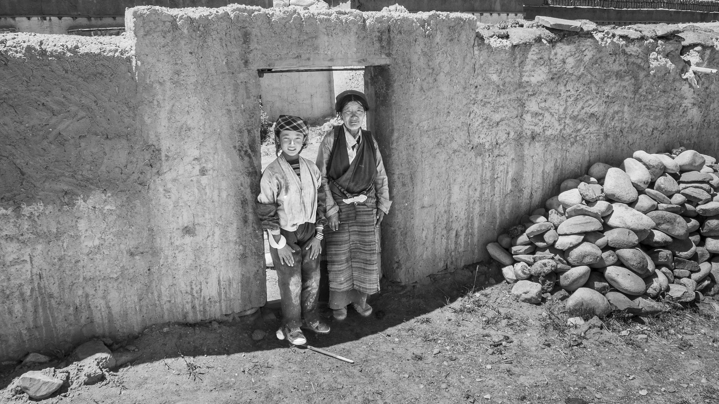 Elderly woman and young girl standing in a doorway of a traditional stone house in rural Tibet.