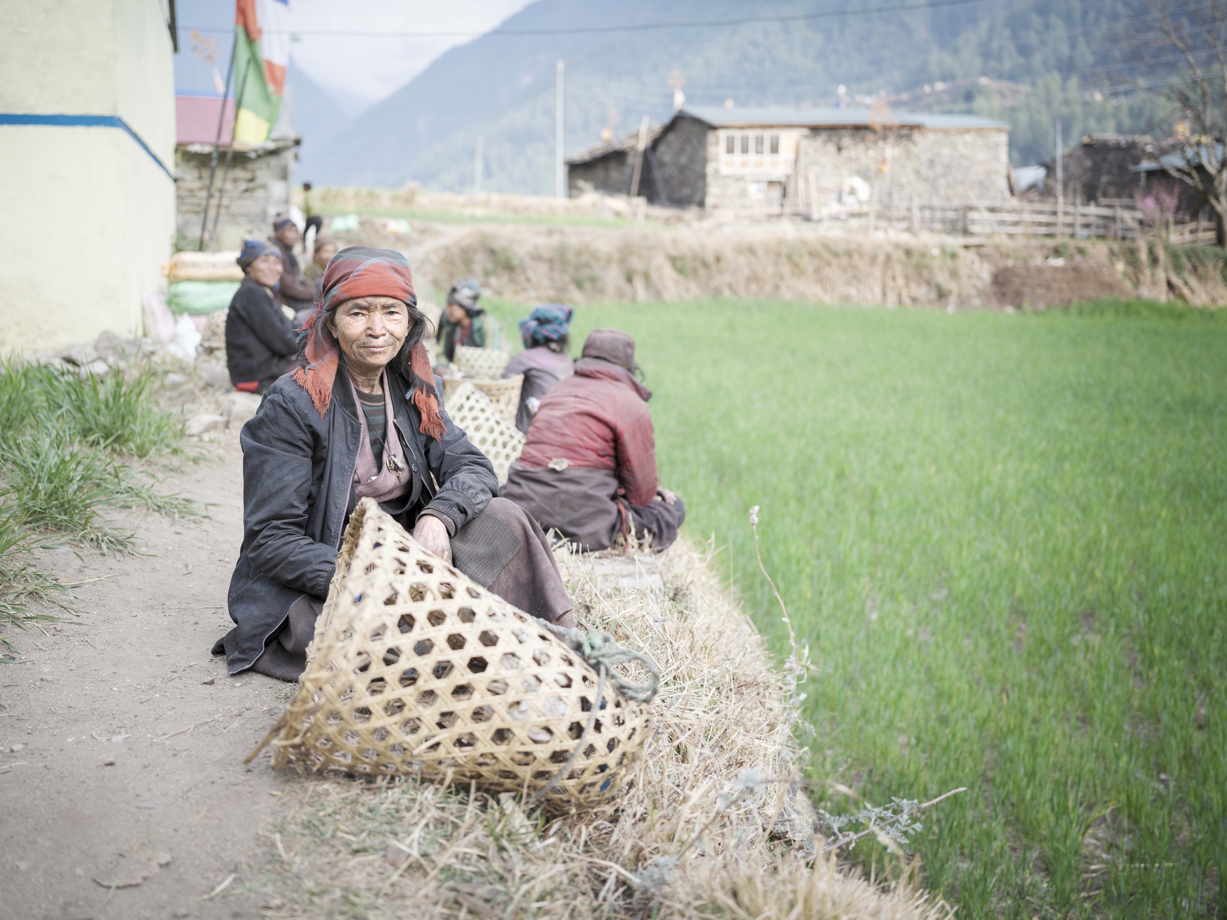 Women pause along the edge of the fields in Tsum Valley, baskets set down beside them. Their days move between tending crops, herding animals, and breaking stones for construction. Rest comes briefly, shared in conversation, before work resumes. Tsum