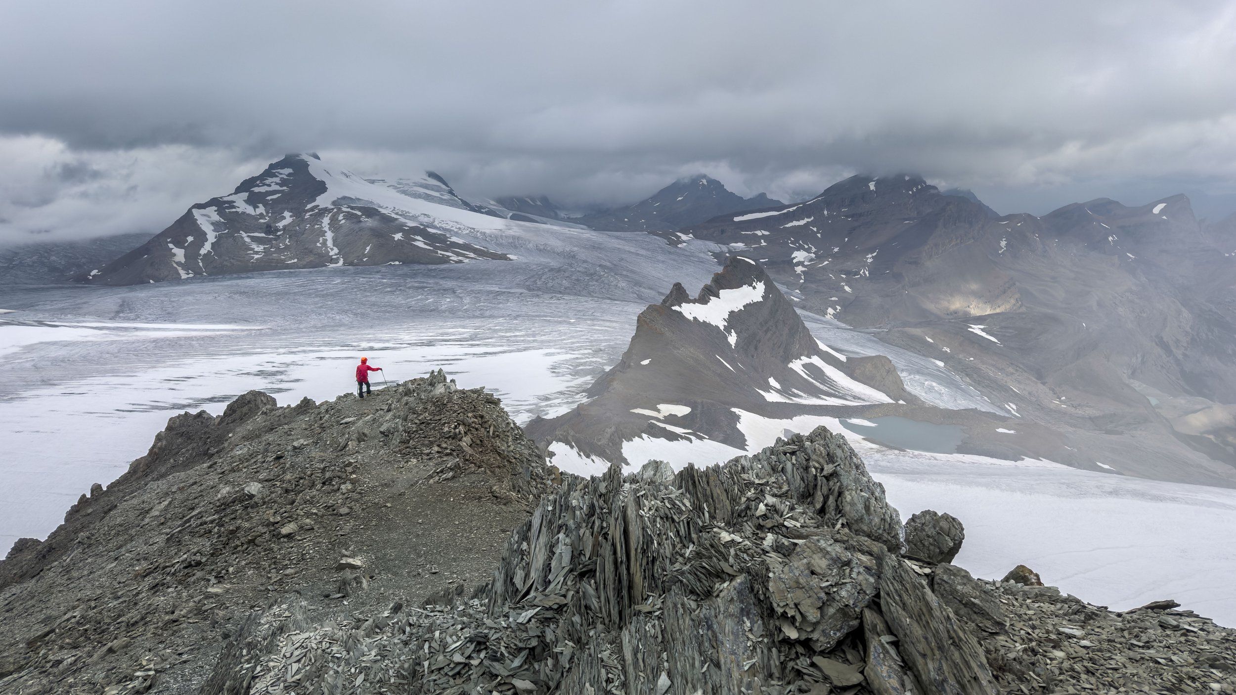 A person in a red jacket standing on a rugged rocky ridgeline in a mountainous, snowy landscape with glaciers and snow-capped peaks under a cloudy sky.