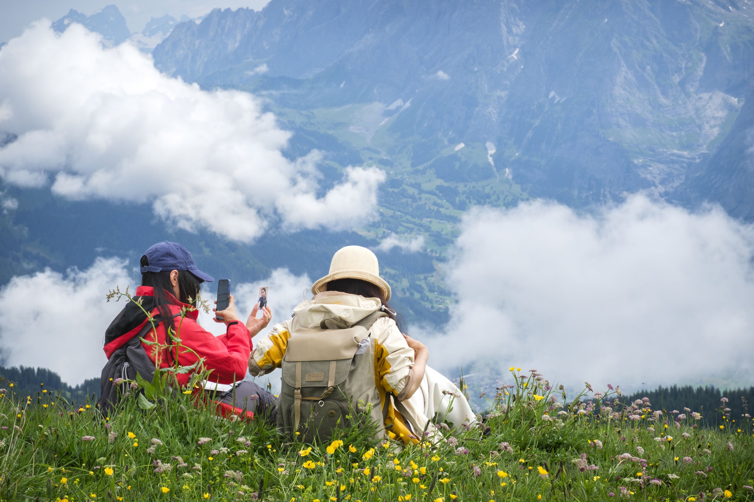 Two women sitting on a grassy field with wildflowers, taking photos of the mountain scenery with clouds and distant mountains in the background.